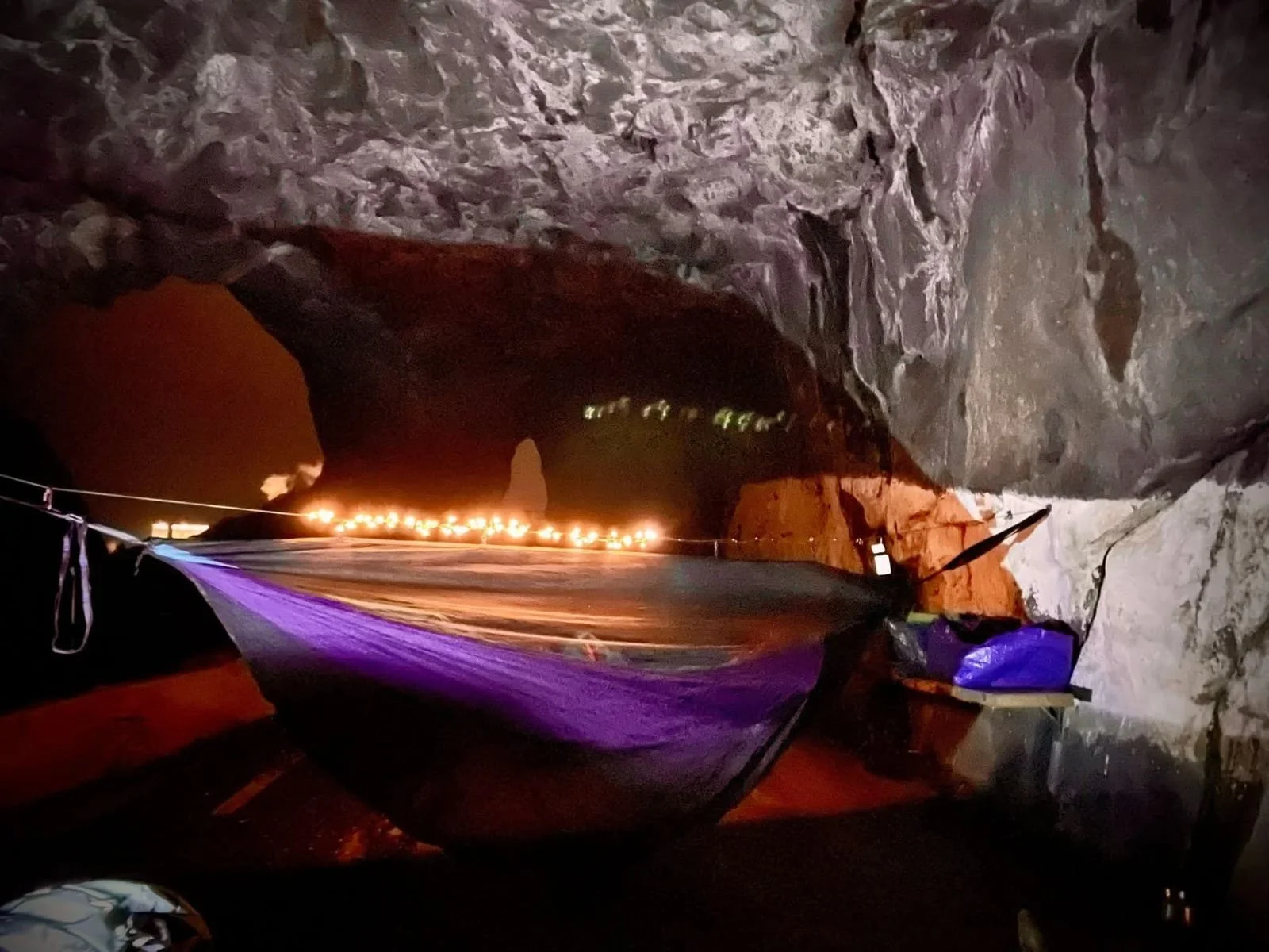 Night hammock setup inside Tunnelstuo tunnel, lit by firelight and string lights against rugged stone walls in Flekkefjord