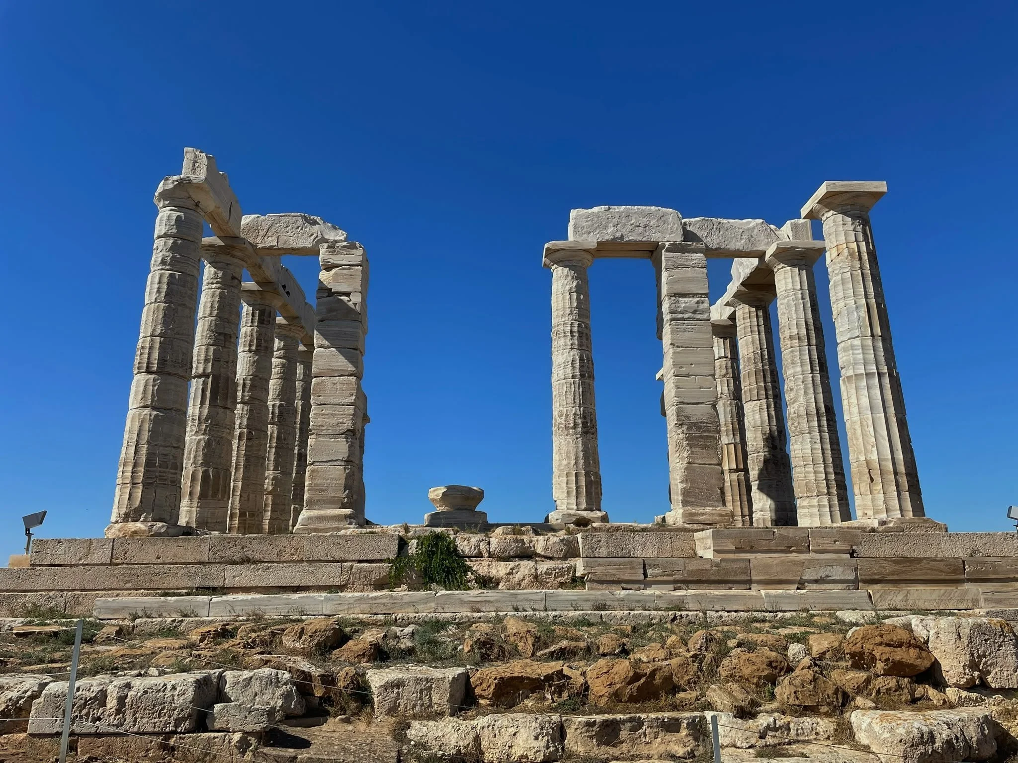 Weathered marble columns of the Temple of Poseidon at Cape Sounion, Greece, rising above stone steps against a clear blue sky.