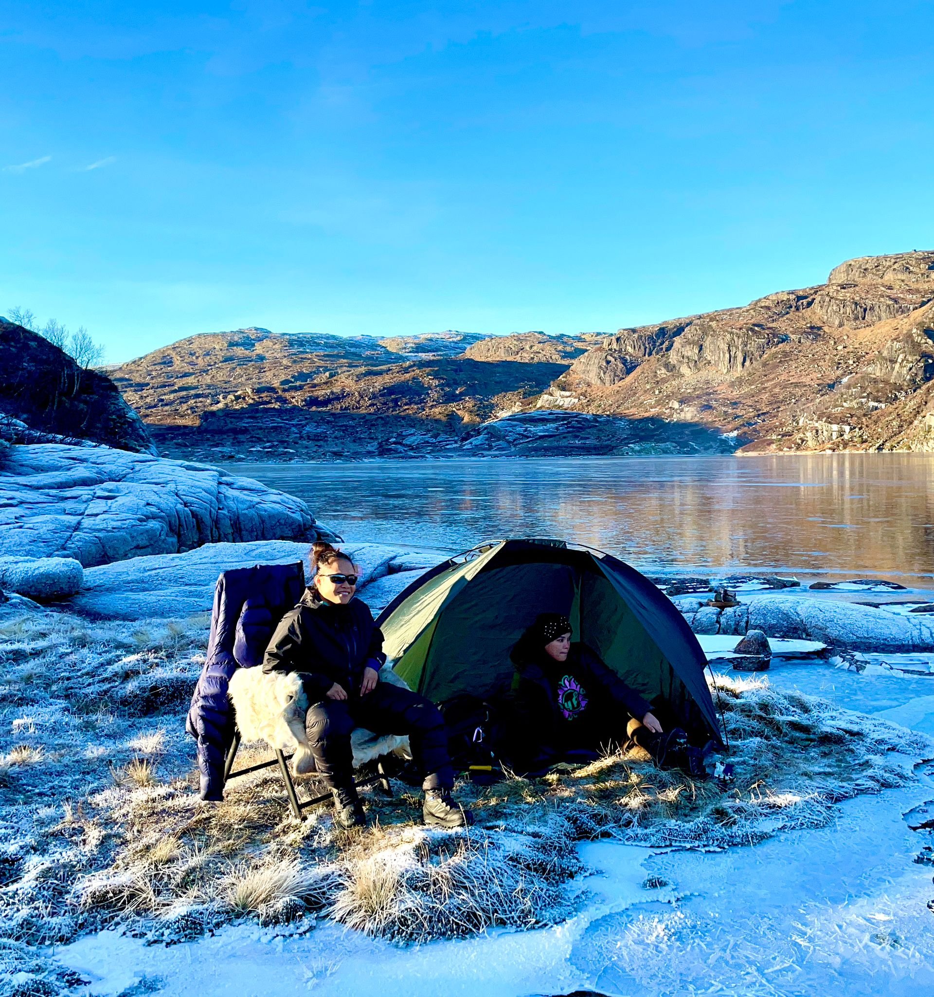 Two people sitting beside a tent at the edge of a frozen Norwegian lake.
