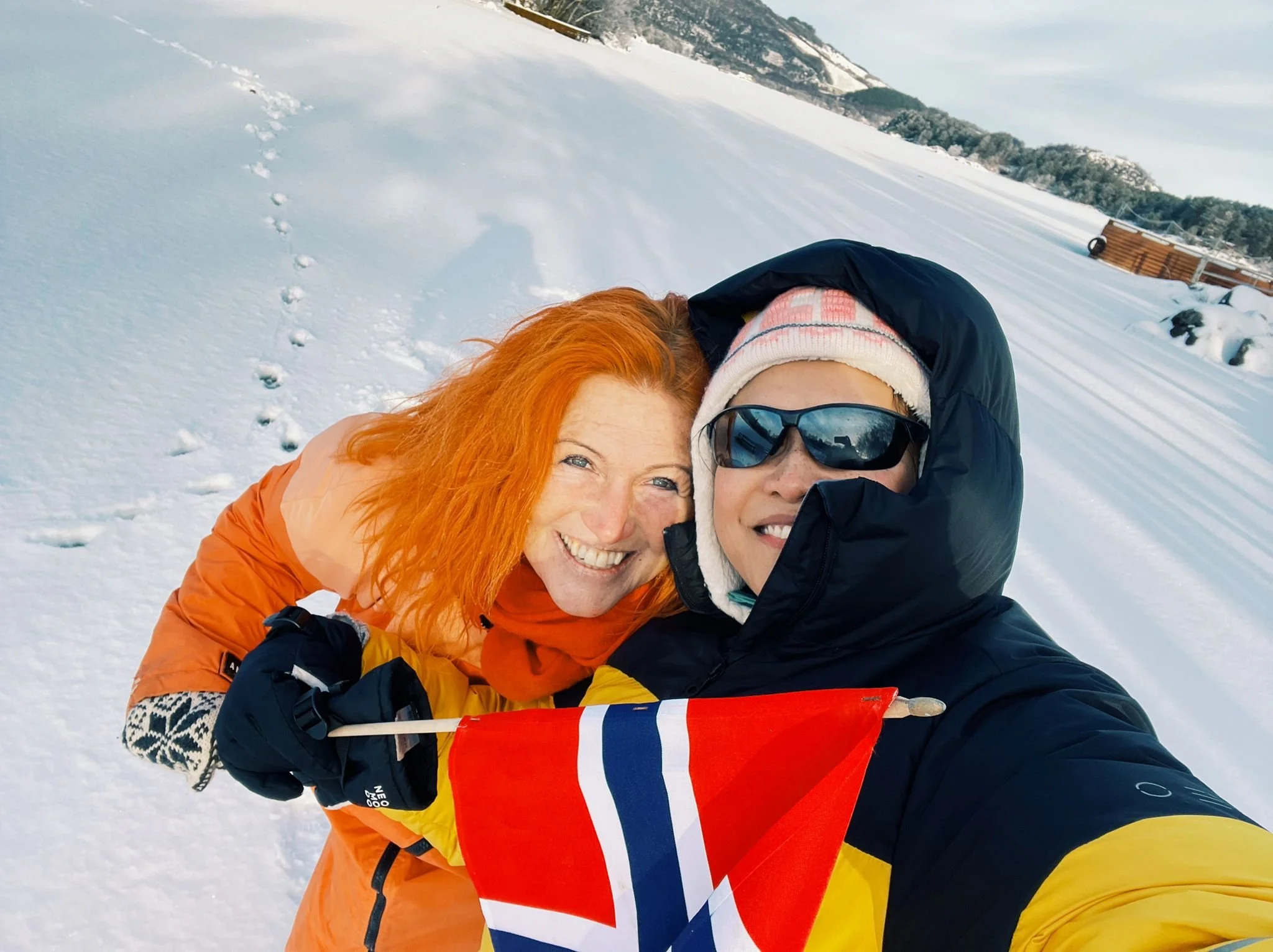 Solveig Fredriksen and April Joy Alfarnes after ice bathing at Alsvik frozen lake in Rogaland, Norway, holding a Norwegian flag during winter cold exposure training.