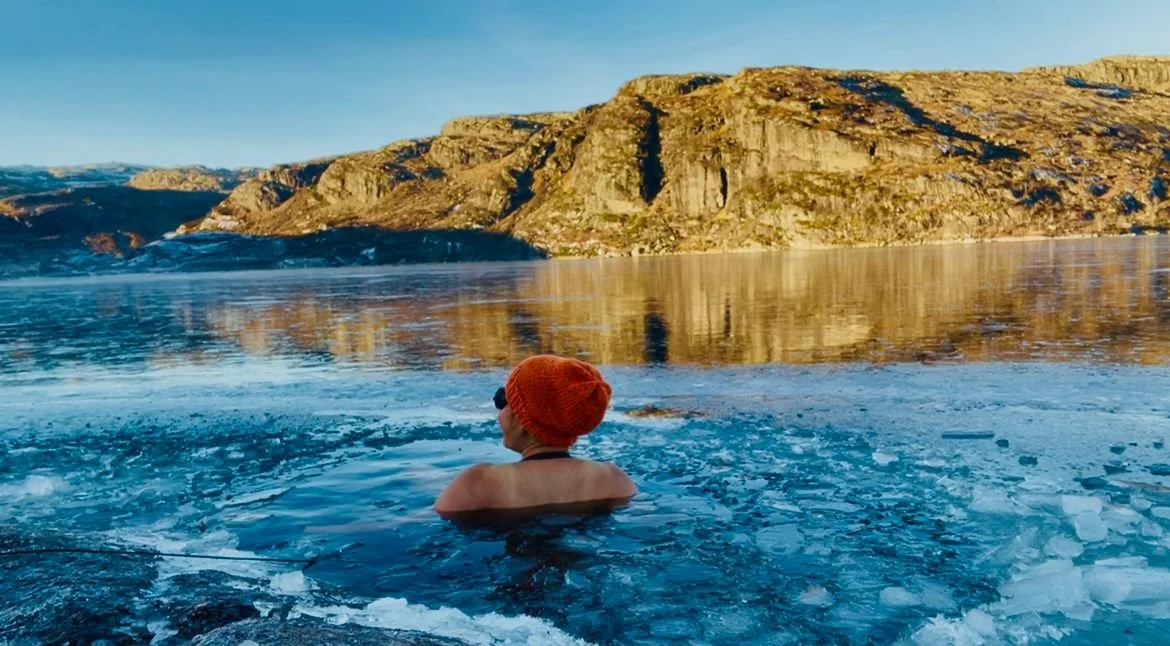 April Joy Alfarnes cold-plunging in a frozen lake in Norway, winter mountains reflecting on the ice under bright blue sky.