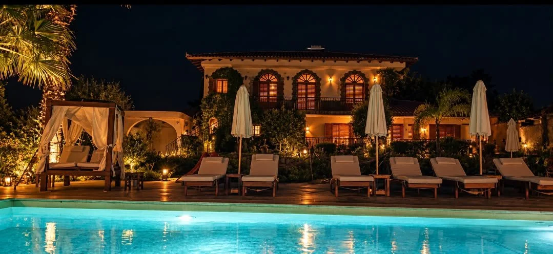 Villa Artemida in Vravrona at night, with warmly lit arched windows, poolside loungers, and a glowing turquoise swimming pool.