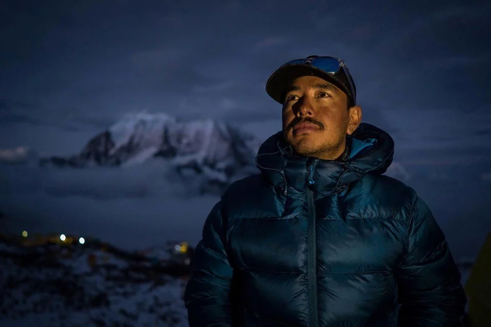 Nirmal "Nimsdai" Purja standing outdoors in a blue down jacket at dusk with snow-covered Himalayan mountains in the background.