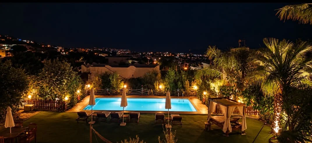 Elevated night view of Villa Artemida’s turquoise-lit pool and palm-lined garden in Vravrona, with distant hillside lights.