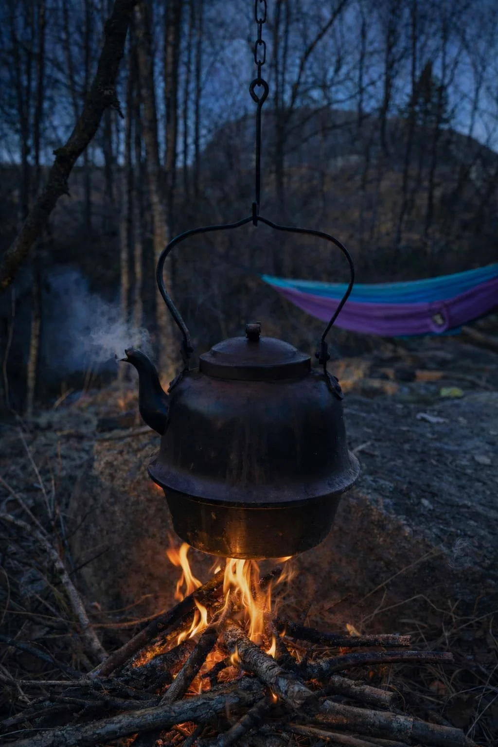 Metal kettle boiling over an open campfire with a purple and turquoise hammock set up in the winter forest at Sviland Falls, Norway.