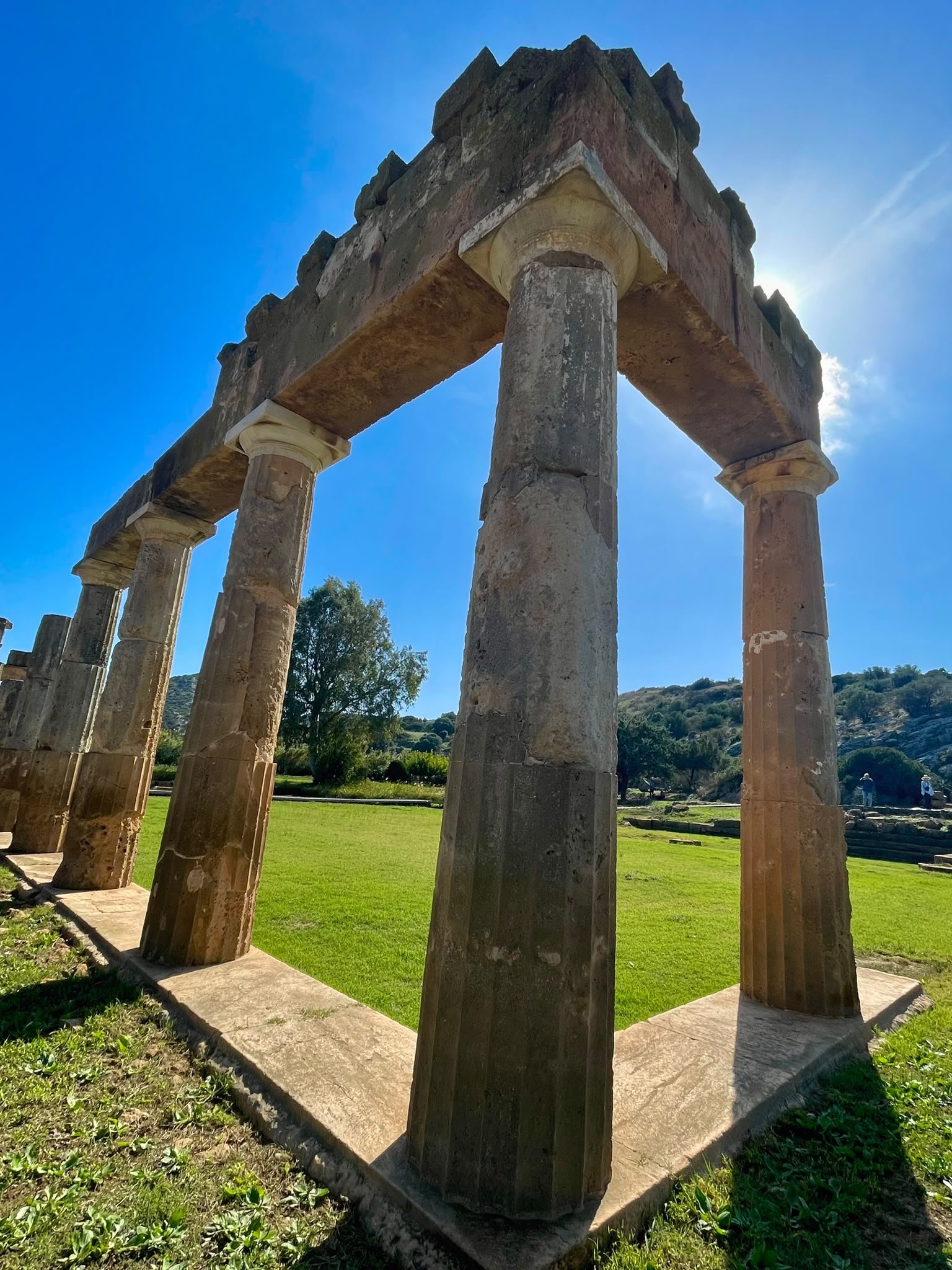 Low-angle view of weathered stone columns and overhead beams at the Sanctuary of Artemis in Brauron, Greece, framing a sunlit green courtyard.
