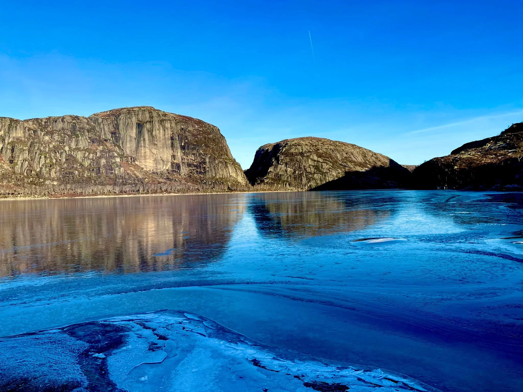 A frozen Norwegian lake reflecting towering mountain cliffs under a blue winter sky.