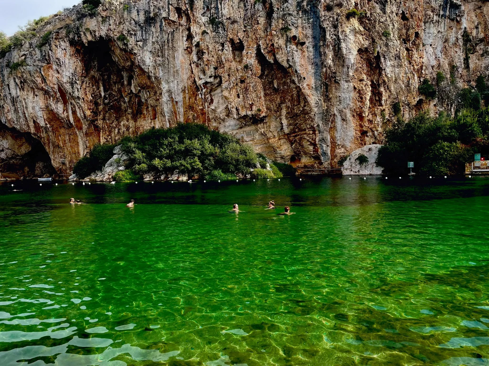 Swimmers in the clear emerald-green water of Lake Vouliagmeni near Athens, Greece, beneath towering, rugged cliffs.