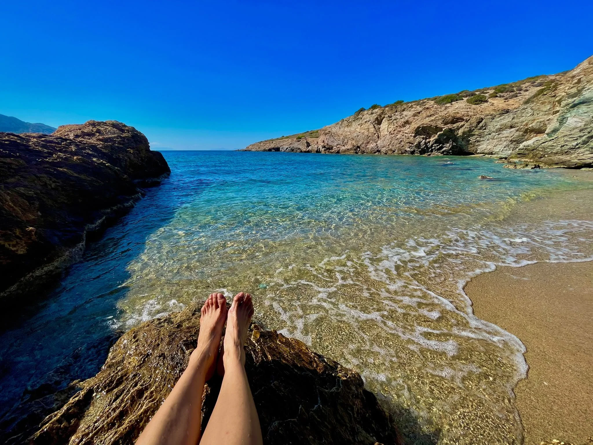 Bare legs resting on a rock above clear turquoise water in a small cove near KAPE Beach, framed by rocky cliffs and a sandy shore.