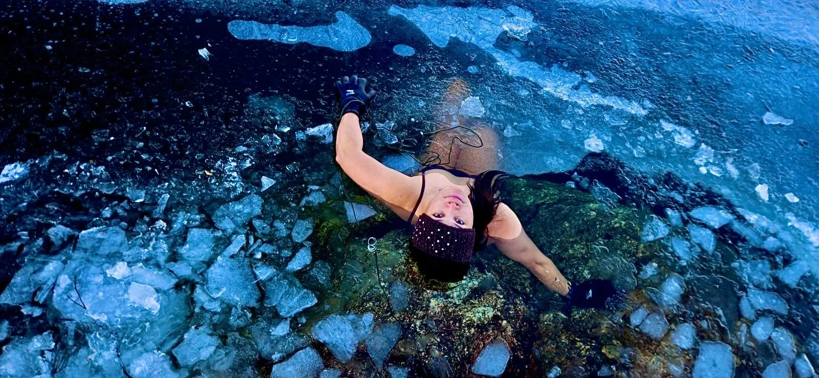 A woman submerged beneath cracked ice, viewed from above in icy blue water