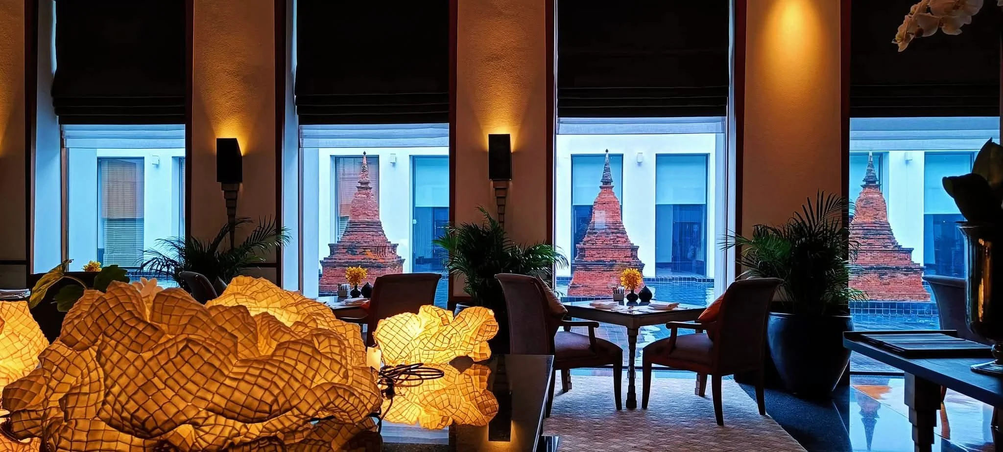Interior of The Sukhothai Hotel in Bangkok featuring warm ambient lighting, elegant lounge seating, and a view of the hotel’s signature brick pagoda structures through large windows