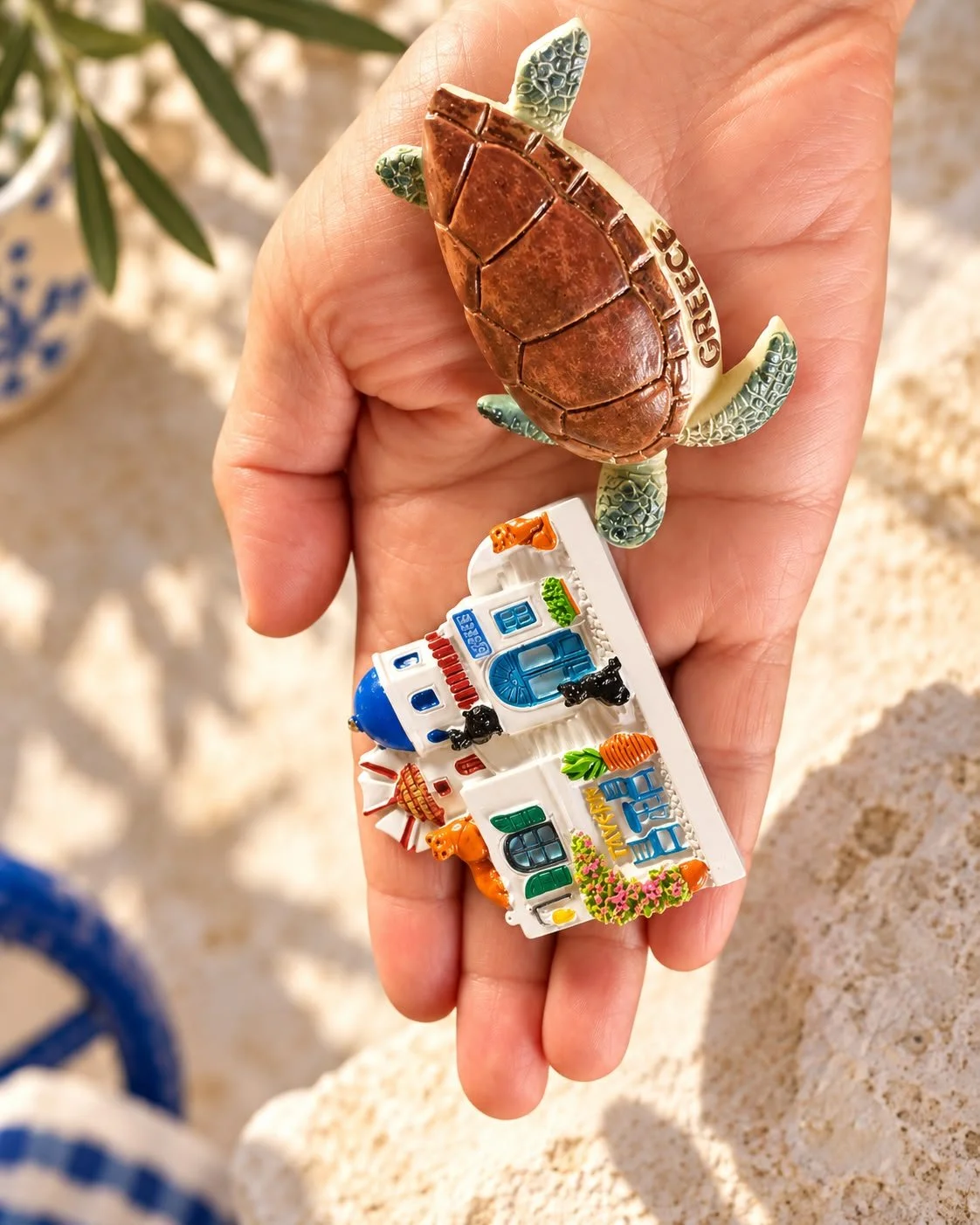 April Joy Alfarnes’s open palm holding two Greek souvenir magnets: a sea turtle marked Greece and a colourful miniature taverna.