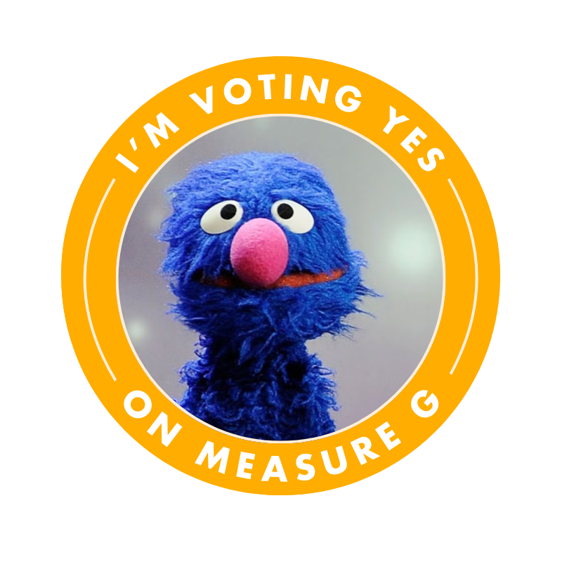 Cotton-headed muppet character with blue fur, pink nose, and white eyes, wearing a yellow campaign badge that says 'I'm Voting Yes on Measure G'.