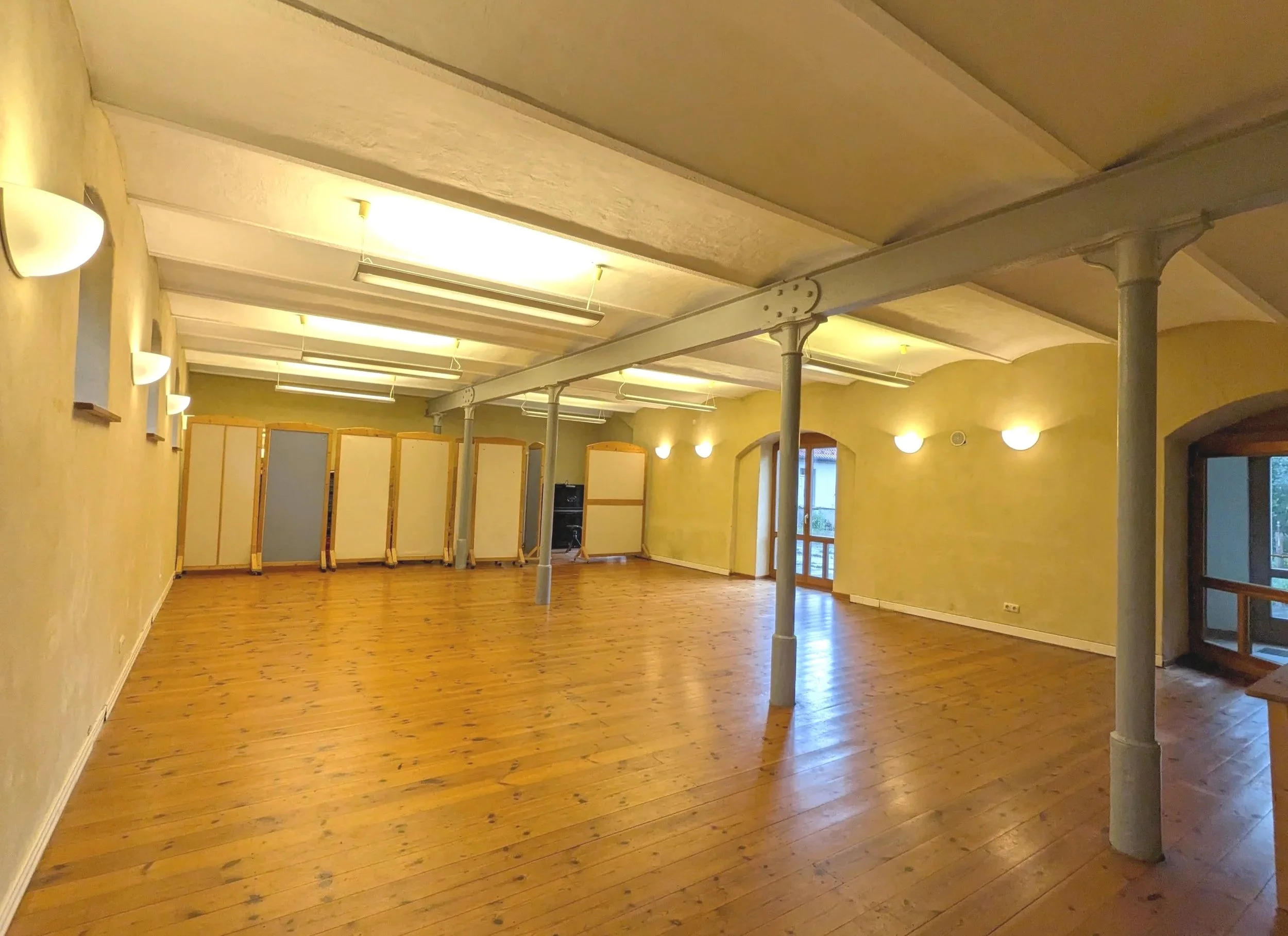 An empty room with wooden floors, yellow walls, and a ceiling supported by columns and beams. There are multiple wall-mounted lights and foldable partition screens in the background.