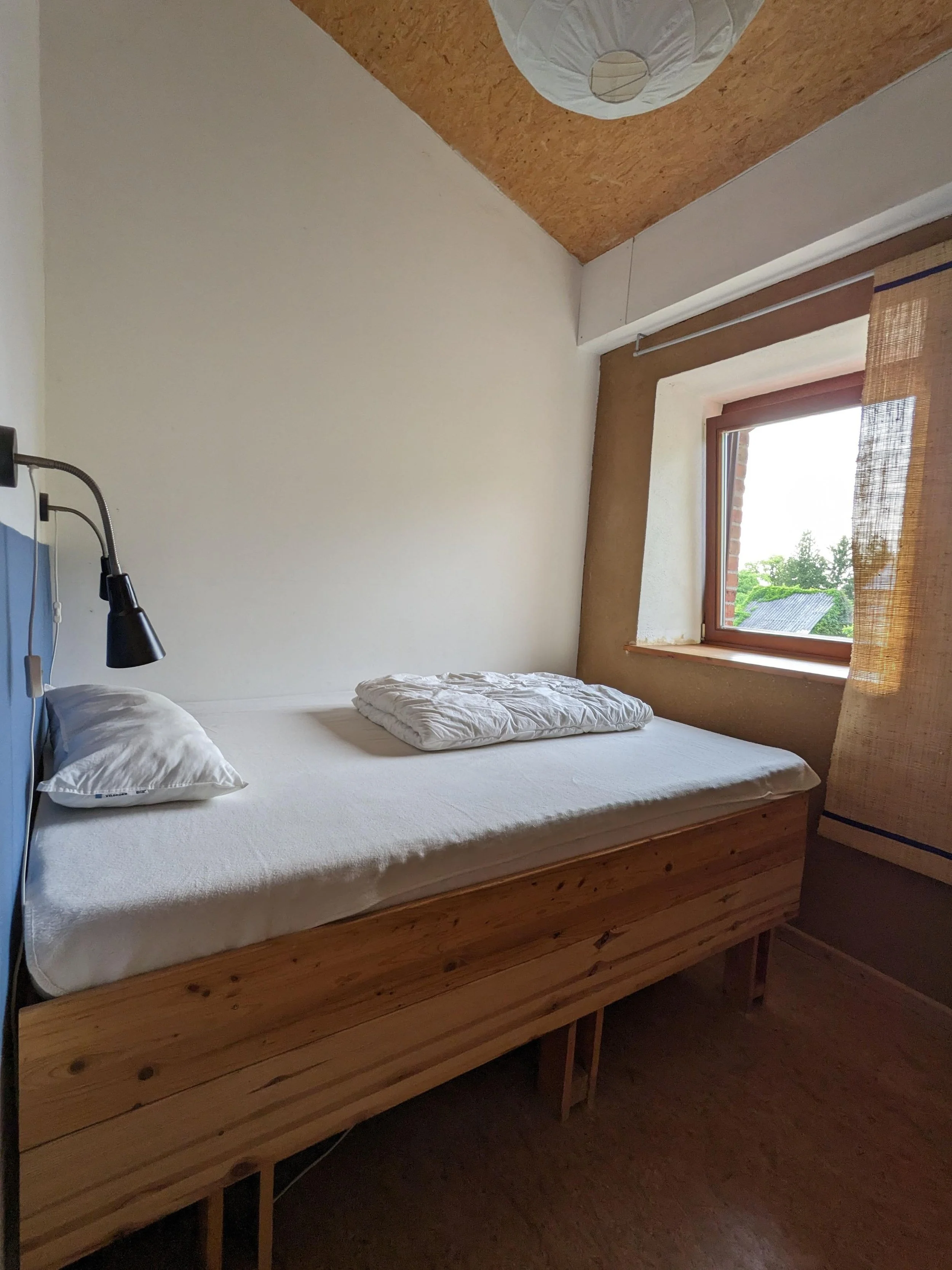 A bedroom with a wooden bed frame, a white mattress, two pillows, a wall-mounted black reading lamp, a window with brown trim and a bamboo curtain, and a slanted ceiling with a paper lantern light fixture.