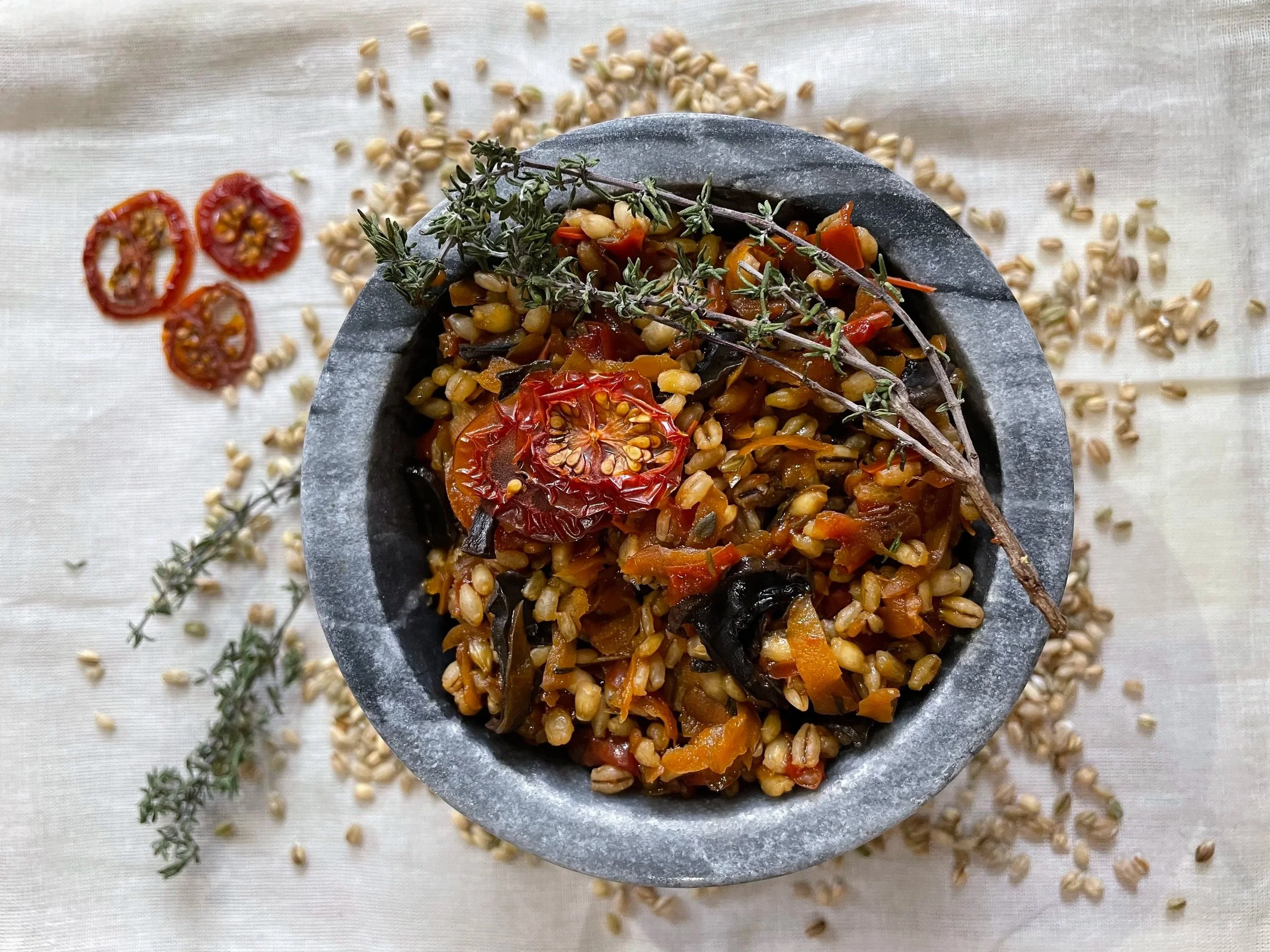 A top-down view of a bowl filled with cooked grains and vegetables, garnished with fresh herbs, surrounded by scattered grains and slices of cherry tomatoes on a light fabric surface.