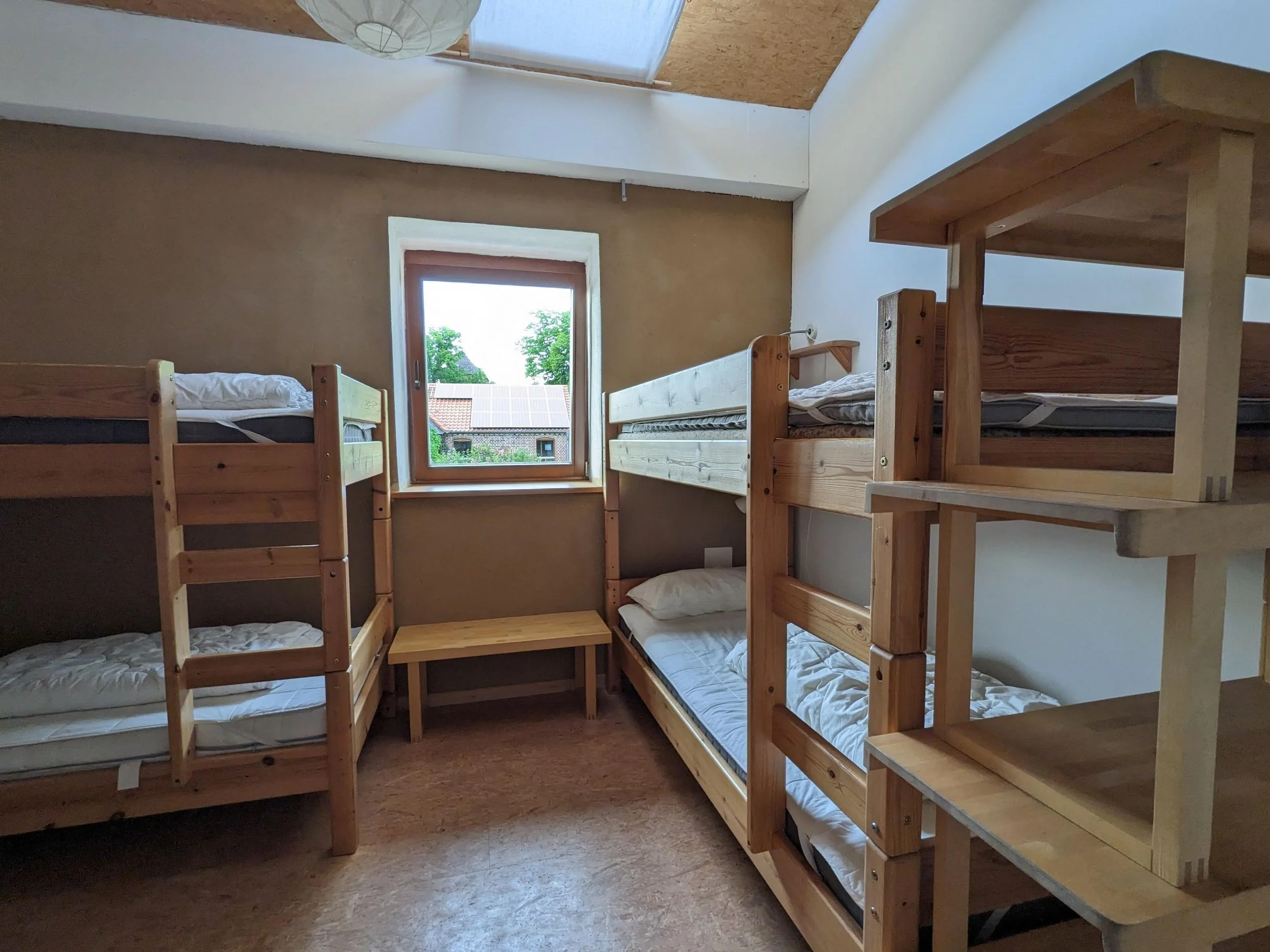 A room with slanted ceiling featuring six bunk beds made of light-colored wood, arranged in two stacks of three, next to a window showing trees and rooftops outside.