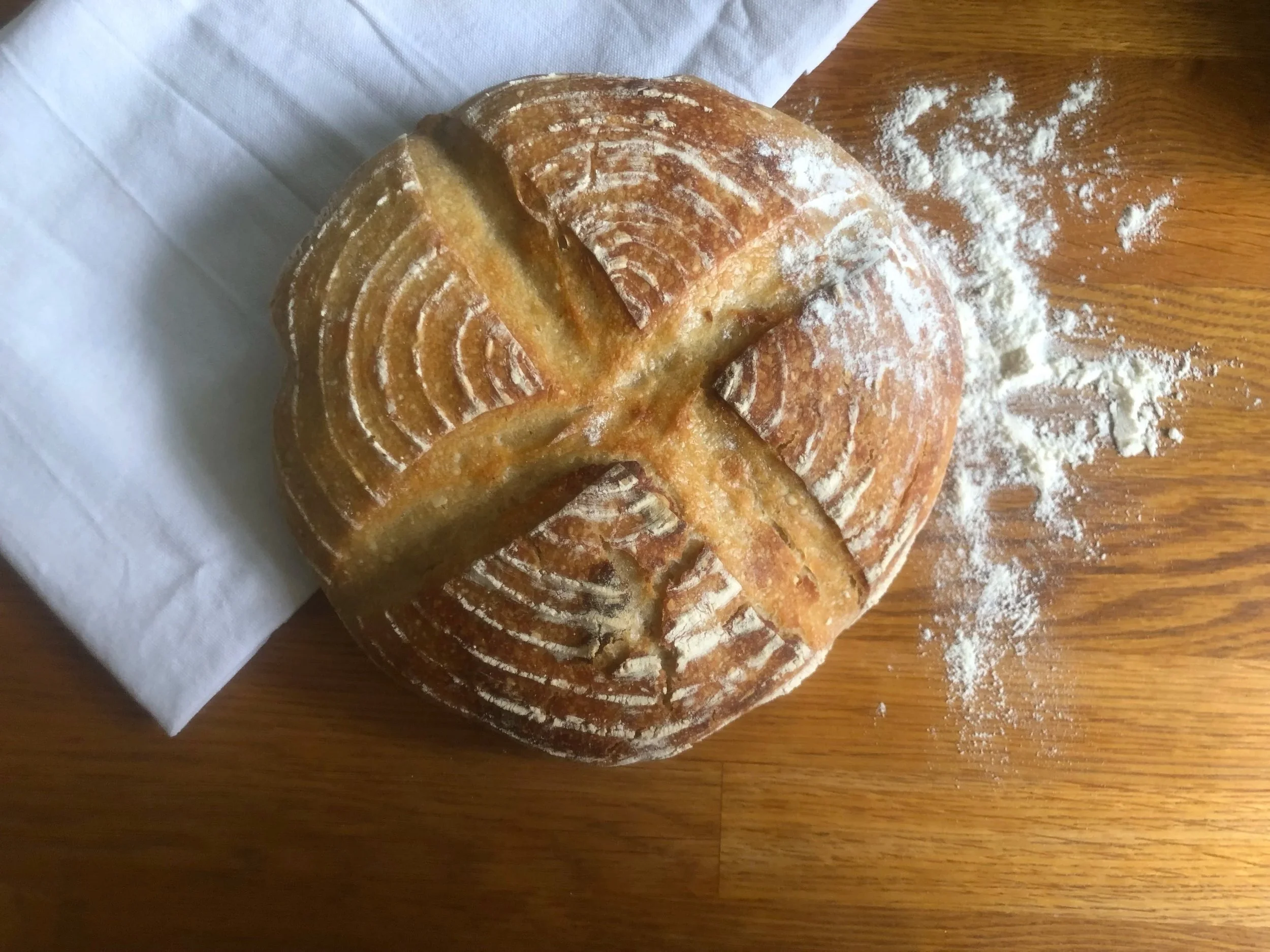 A loaf of bread with a decorative pattern on top, resting on a cloth napkin on a wooden surface with scattered flour.
