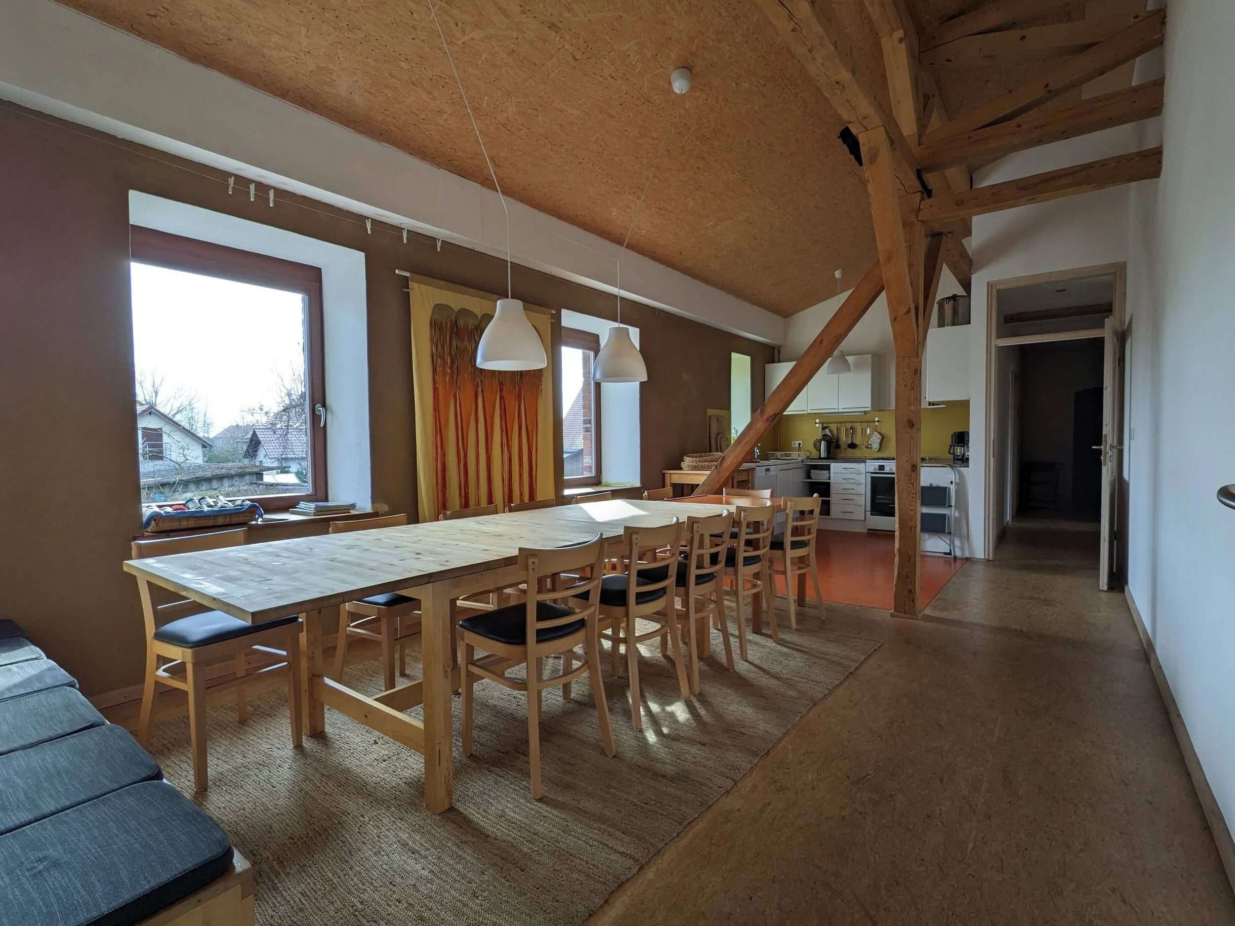 A dining area with a large wooden table and multiple chairs, large windows with view of neighboring houses, a yellow curtain, pendant lighting, and a kitchen in the background with white cabinets and appliances.