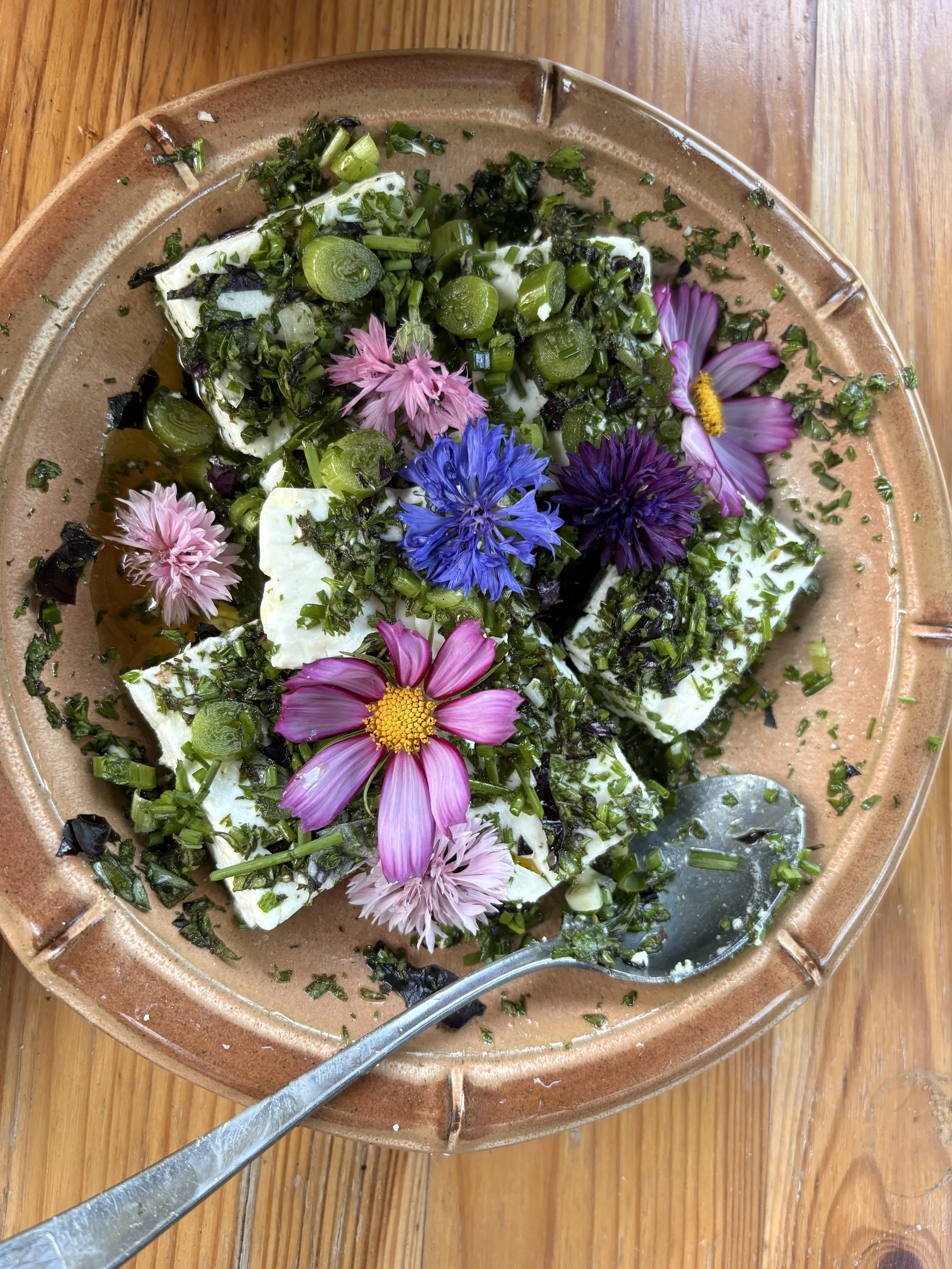 A ceramic plate with sliced cheese topped with chopped green herbs and scallions, garnished with pink, purple, and blue edible flowers, on a wooden table.