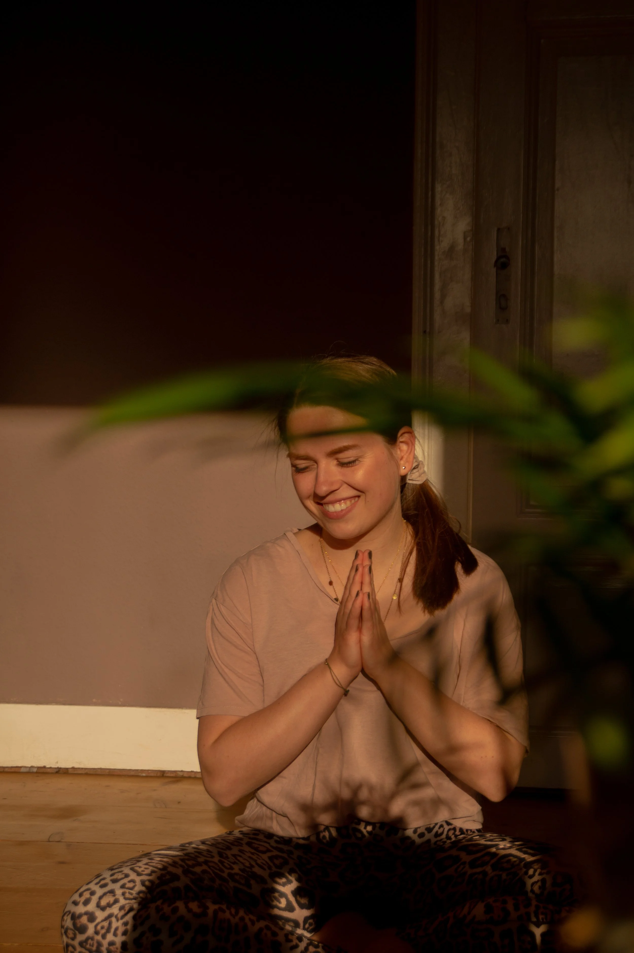 A young woman with brown hair tied back, smiling with her eyes closed, sitting cross-legged on a wooden floor indoors. She has her hands pressed together in a prayer position and is wearing a beige T-shirt and leopard-print pants. The background shows a wooden door and textured wall.