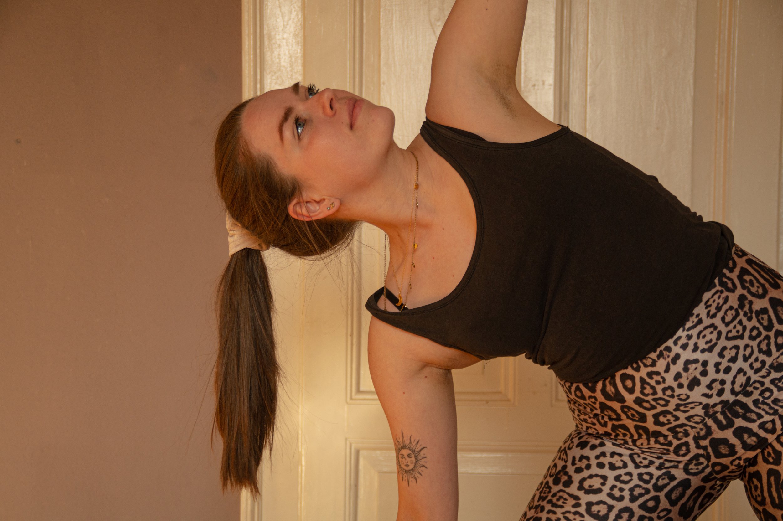 A woman with long brown hair tied in a ponytail, wearing a black sleeveless top and leopard print pants, is performing a yoga pose on the floor. She has a sun tattoo on her forearm, earrings, and a necklace, and she is looking upwards.
