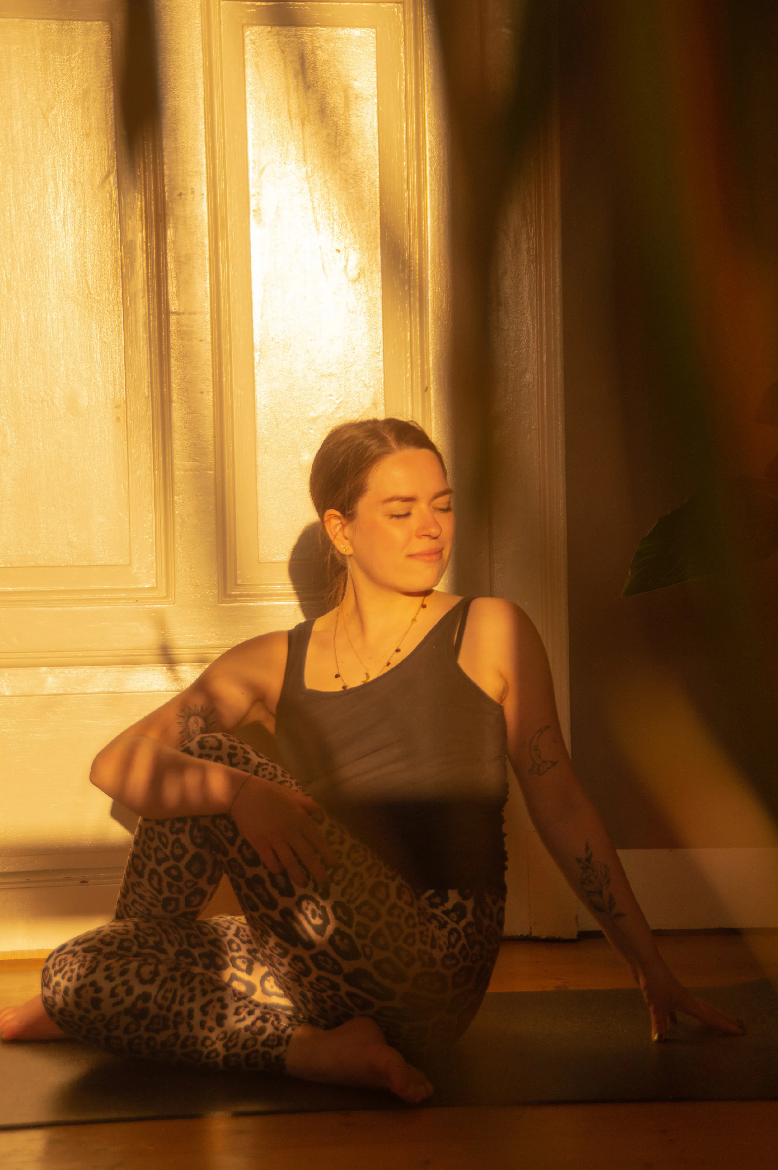 A woman with tattoos on her arms and a leopard print pair of pants, sitting on the floor with her eyes closed, wearing a black tank top, illuminated by warm golden sunlight, in a room with cream-colored walls and decorative molding.