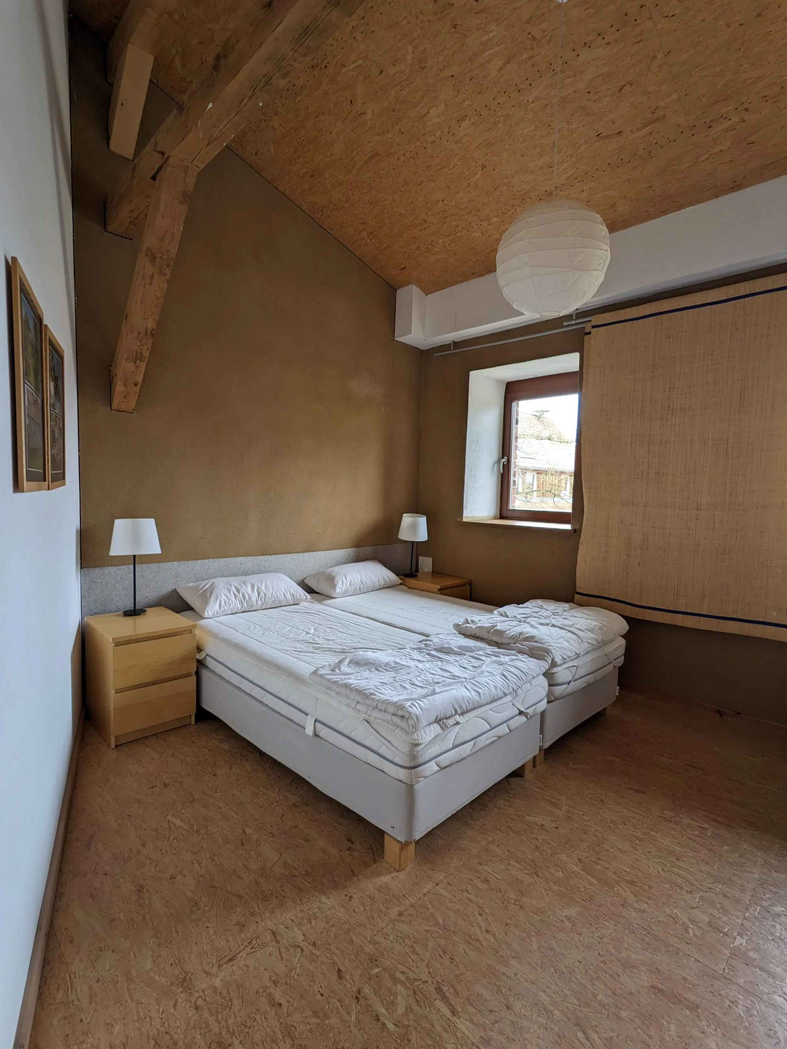 Room with two twin beds pushed together, two nightstands with white lamps, window, hanging paper lantern ceiling light, and wooden ceiling beams.