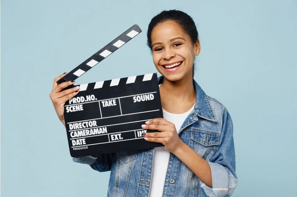 Young woman smiling and holding a film clapperboard against a light blue background.