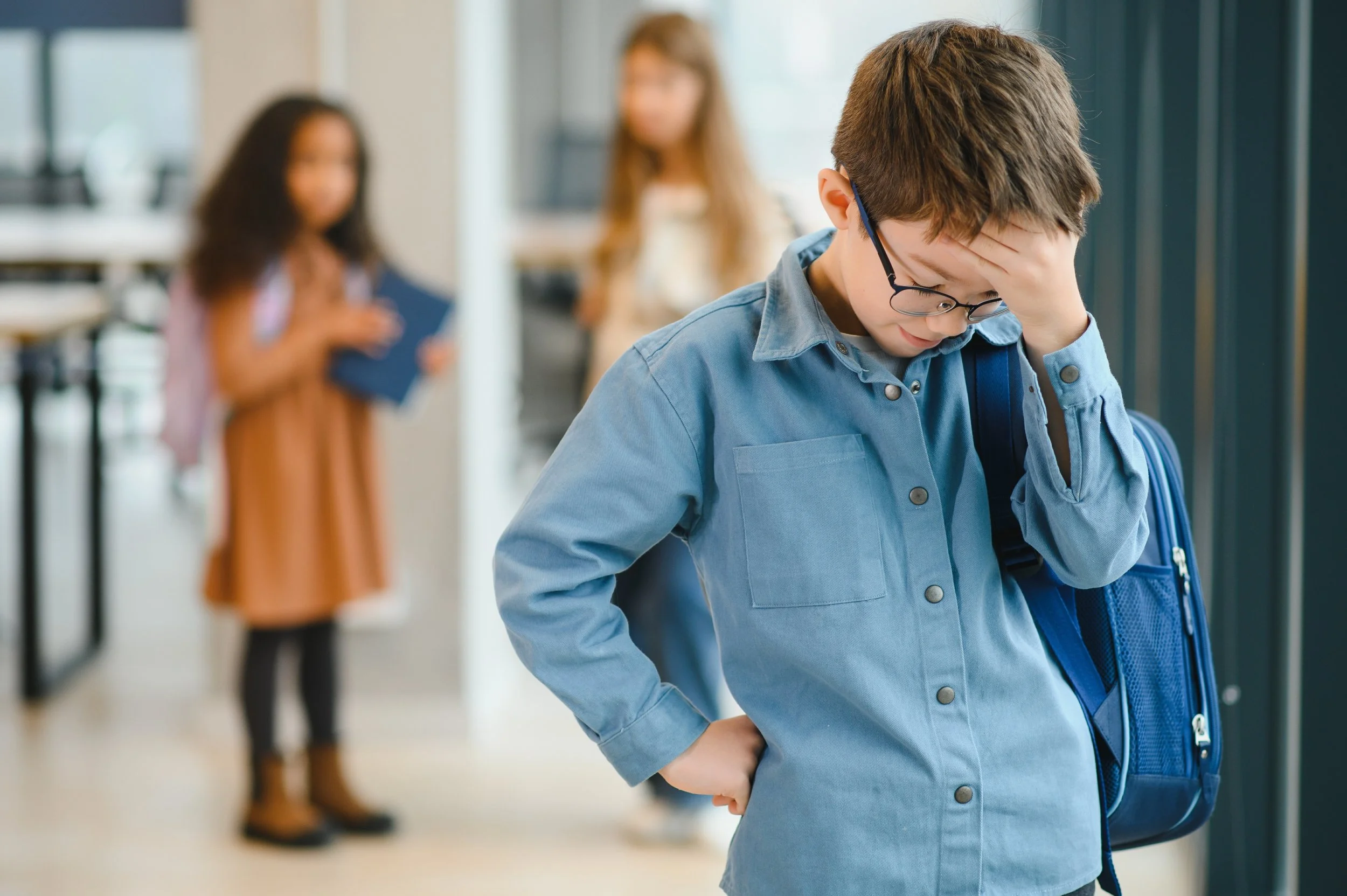 A young boy with glasses and a blue shirt holding his head in frustration or worry, standing in a school hallway with two blurred girls in the background.