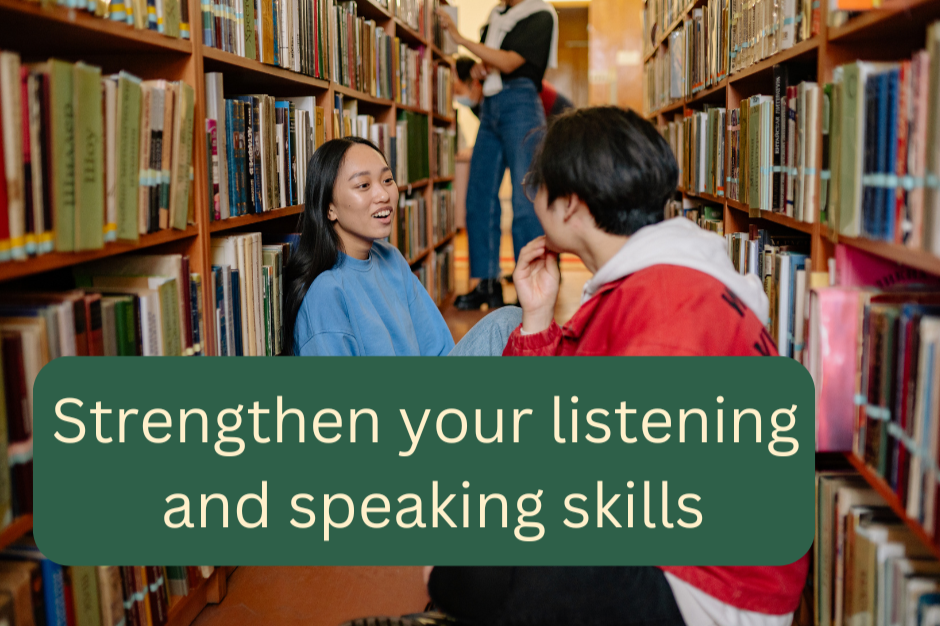 Two young people having a conversation on the floor in a library aisle between two shelves full of books, with a third person standing in the background browsing books.