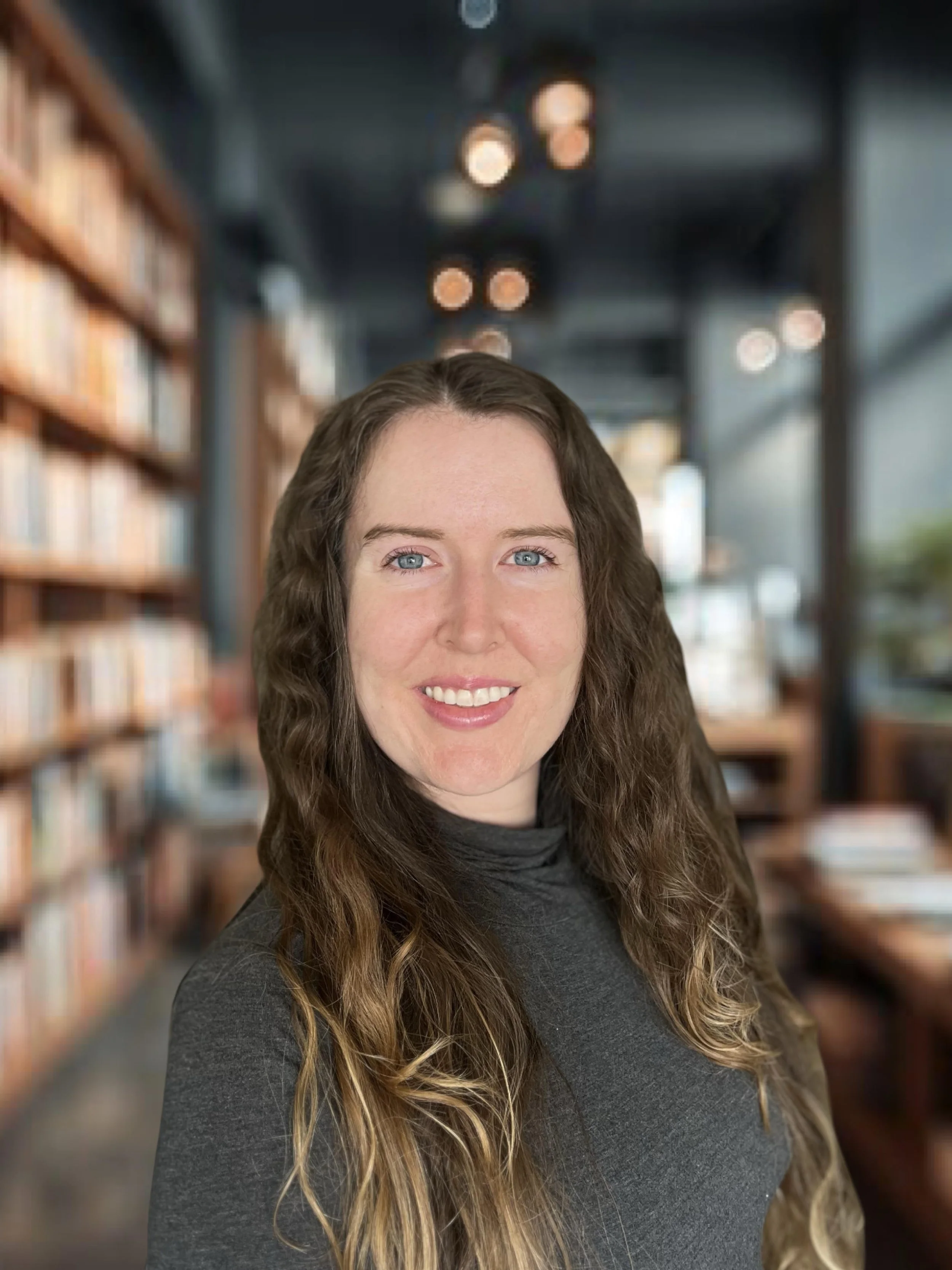 A woman with long, wavy brown hair and blue eyes smiles at the camera in a cozy library setting.