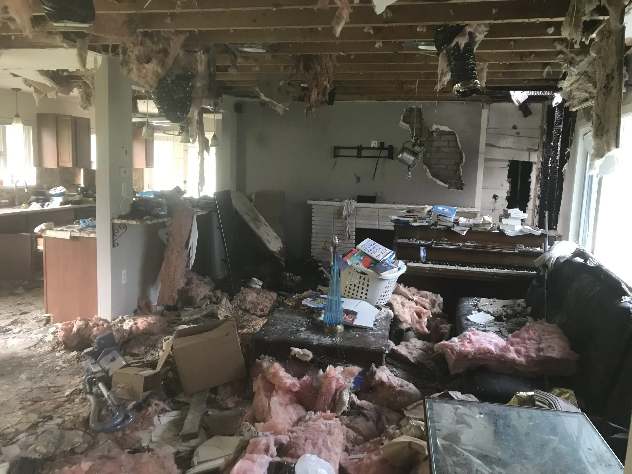 Interior of damaged living room and kitchen with debris, exposed insulation, and disarrayed furniture.