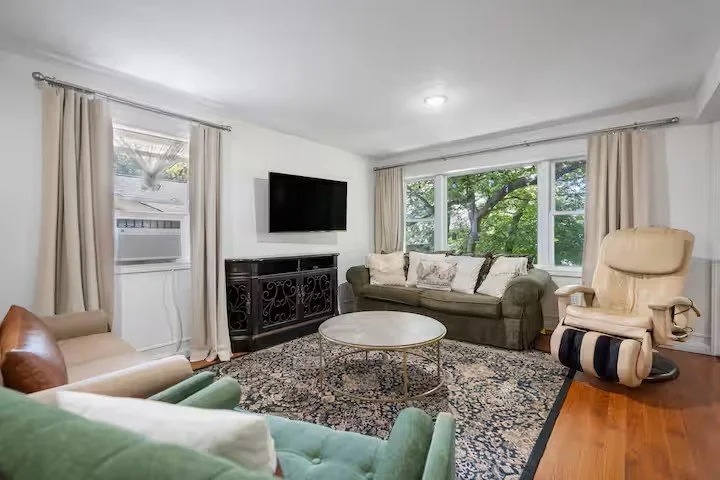 Modern living room with green and beige sofas, round coffee table, patterned rug, wall-mounted TV, and bookshelves. The room has hardwood floors and neutral walls.