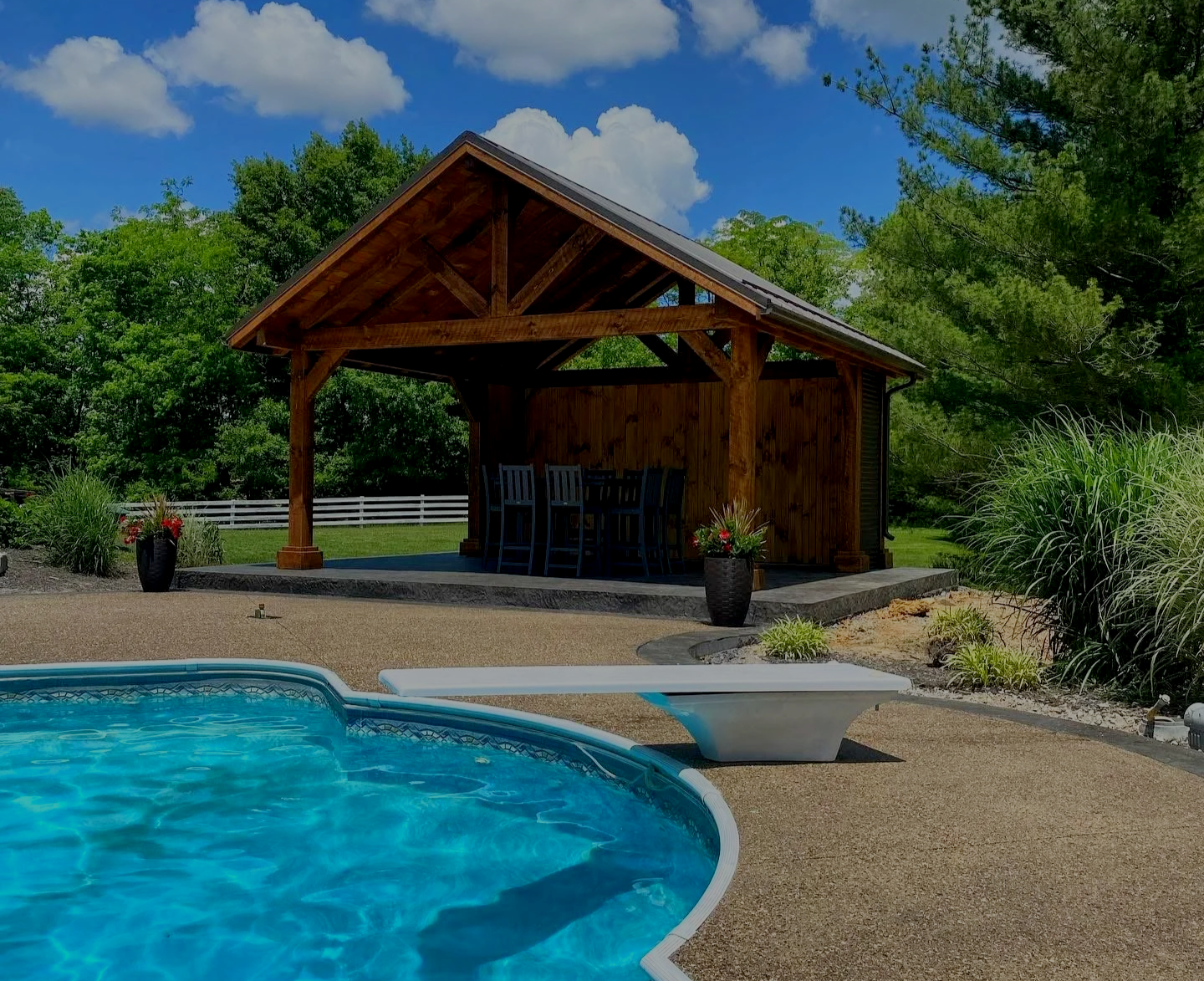 Swimming pool with a diving board in front of a wooden pool house surrounded by trees and blue sky.