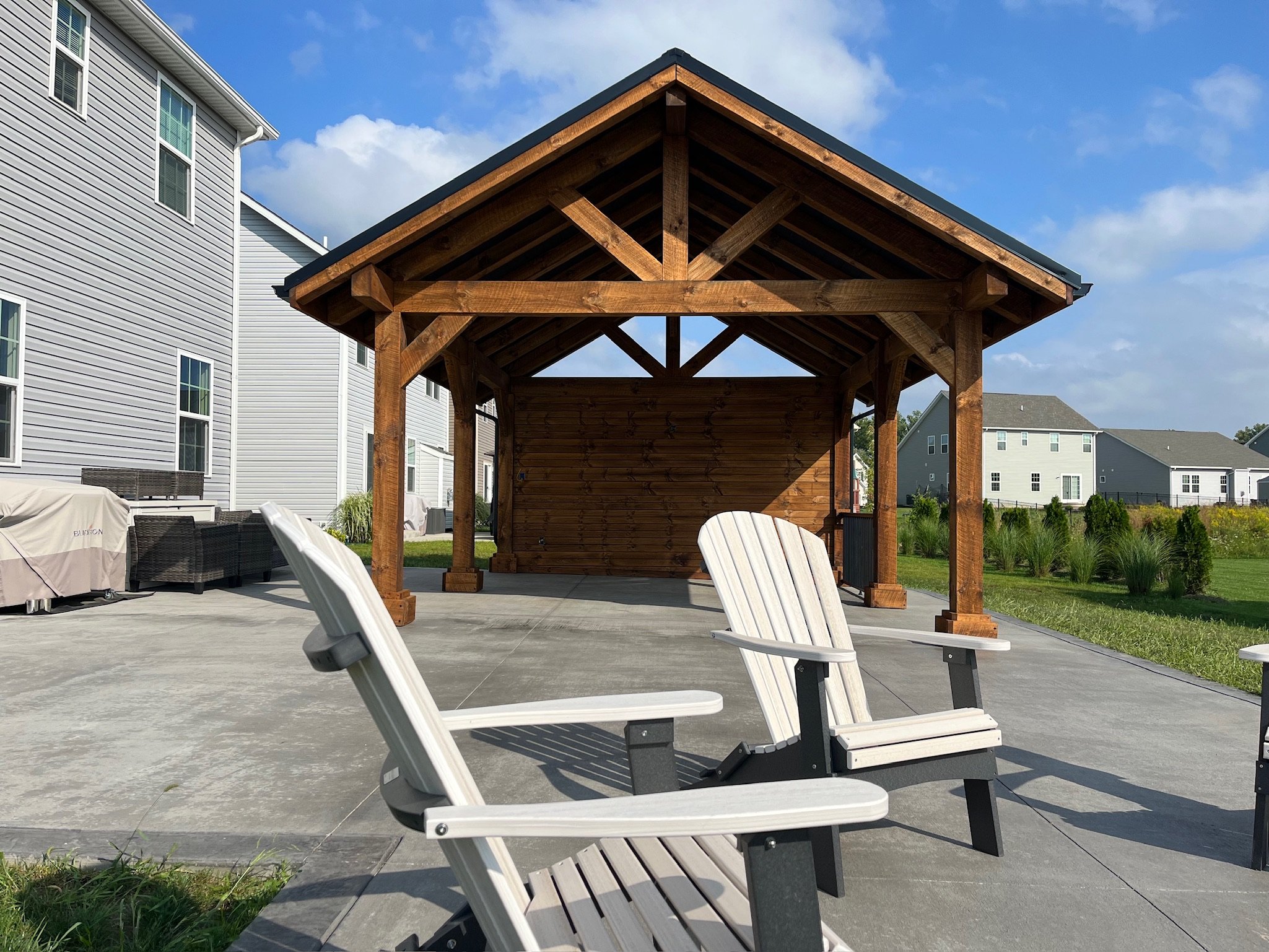 Backyard patio area with two white Adirondack chairs, a wooden gazebo with a peaked roof, and neighboring houses under a partly cloudy sky.