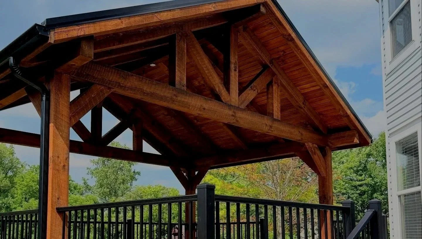 Backyard wooden deck with a covered pergola, surrounded by black metal railing, with trees and part of a white house visible in the background.