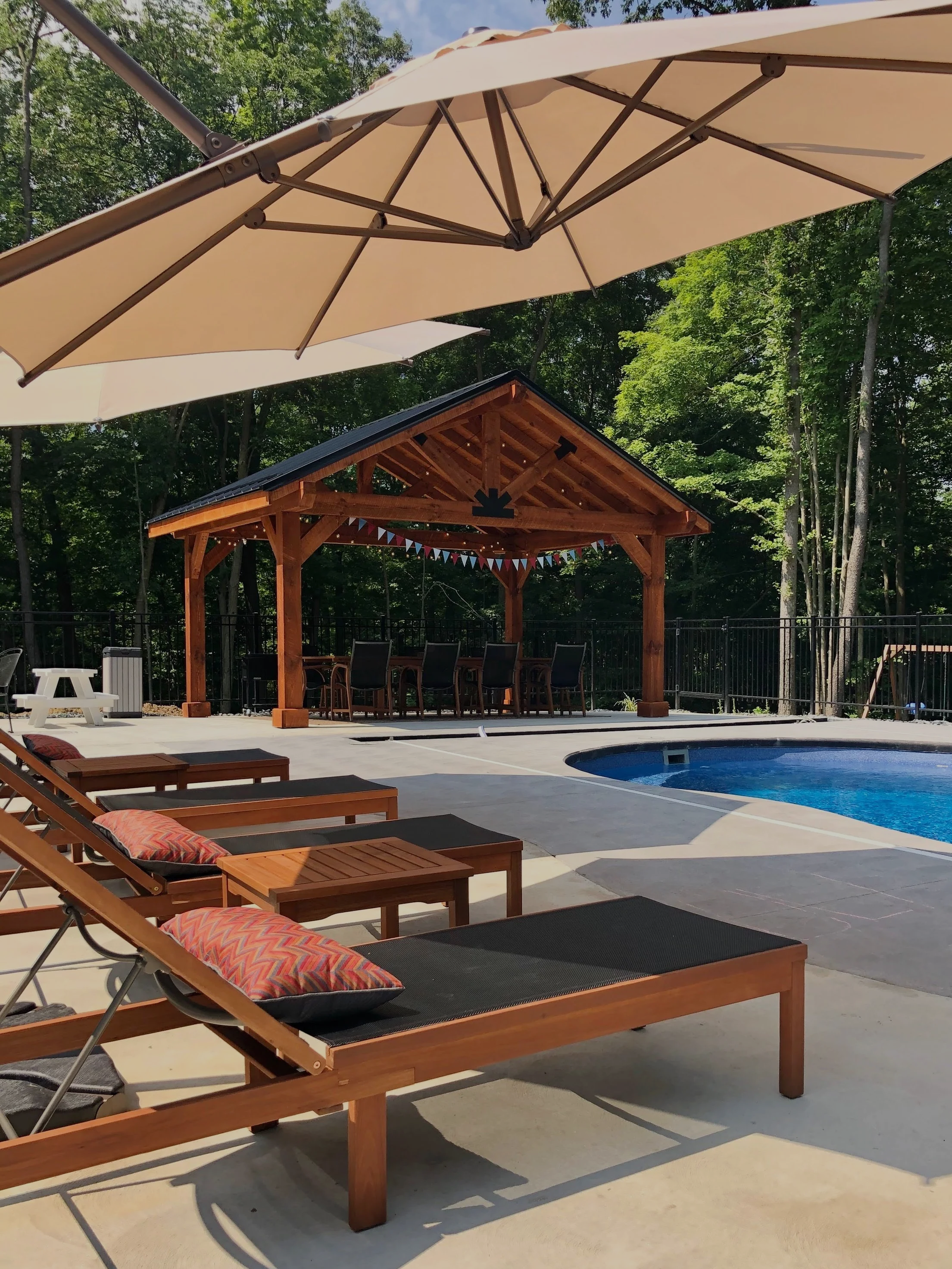 Poolside area with lounge chairs, umbrella, a wooden pavilion with a dining table, surrounded by a forested background.