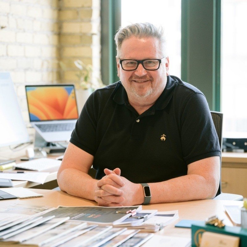 A smiling man with gray hair and glasses sitting at a desk in an office, with a laptop and various papers scattered on the desk, and a window in the background.