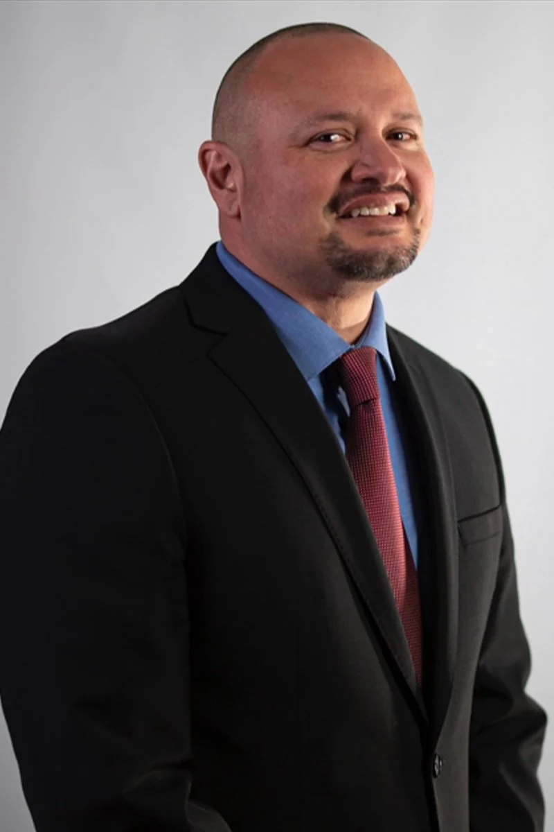 A man in a black suit, blue shirt, and red tie smiling at the camera against a plain light grey background.