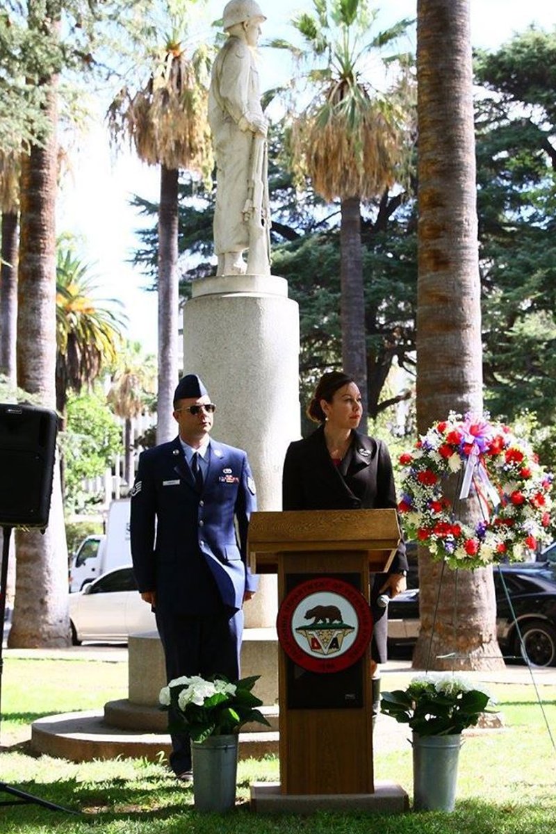 A memorial ceremony outdoors with a statue of a soldier on a pedestal, two people standing at a podium with a wreath of red, white, and purple flowers, and surrounded by palm trees.