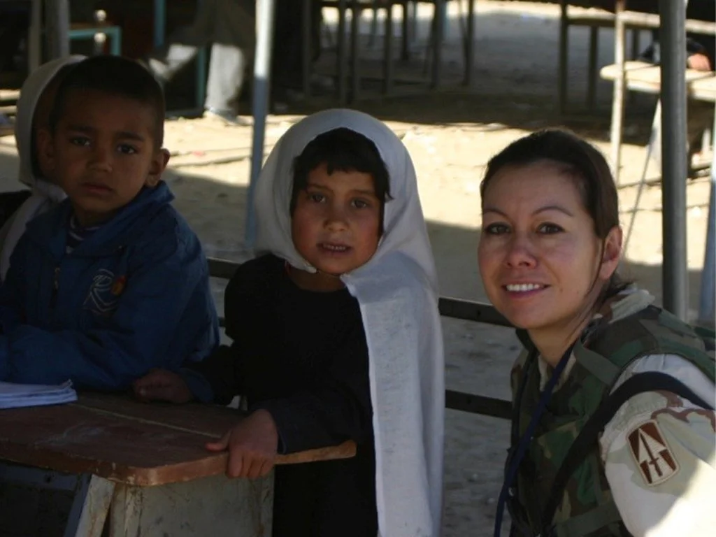 A woman in military uniform and two young boys sitting at a table outdoors, with a school setting in the background.