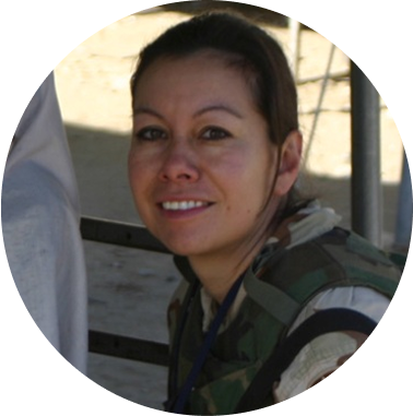 A woman in military camouflage uniform smiling outdoors in a sunny environment.
