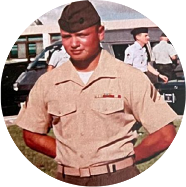 A young man in a beige military uniform and cap standing outdoors with other people and vehicles in the background.