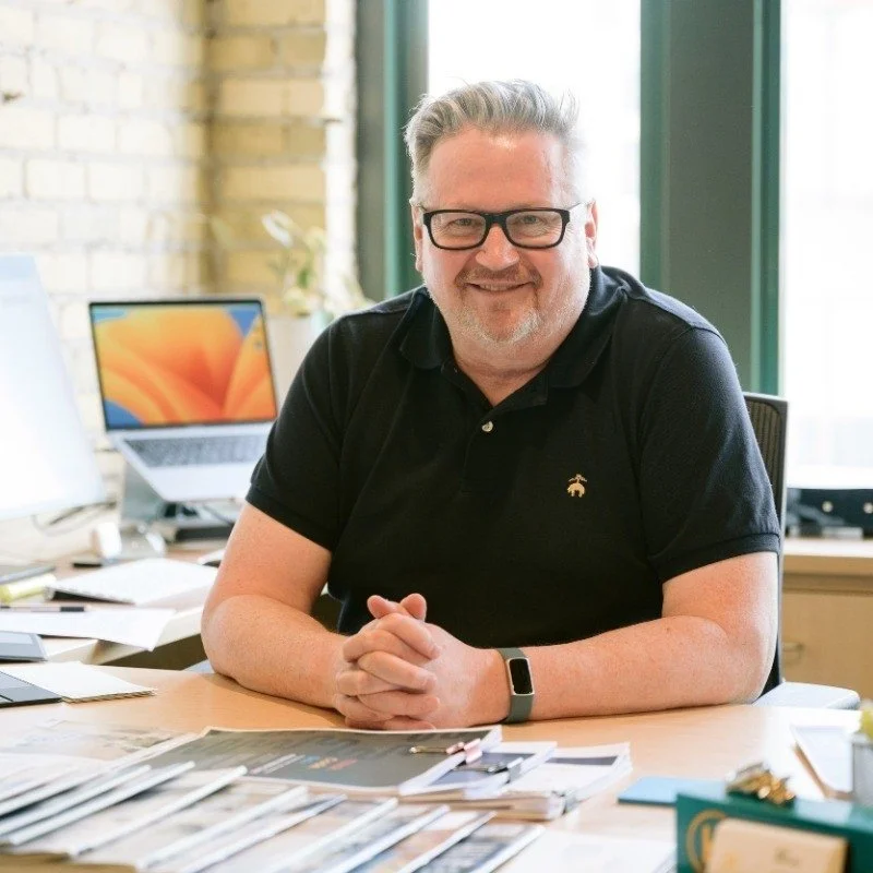 A man with gray hair and glasses smiling and sitting at a desk in an office with a laptop and papers.