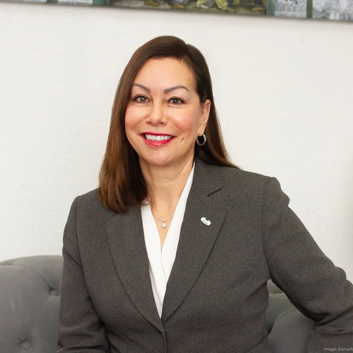 A woman with brown hair, wearing a dark gray blazer and a white shirt, smiling while seated on a gray sofa, against a white wall with a shelf and posters above.