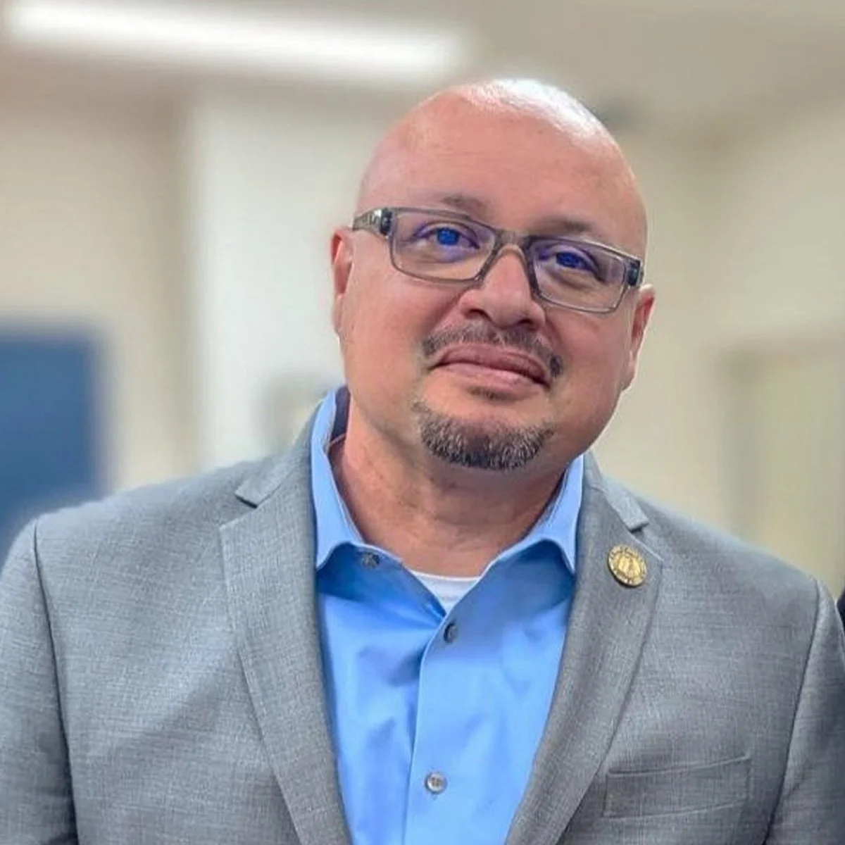 A man wearing glasses, a light gray suit, and a blue shirt, smiling.