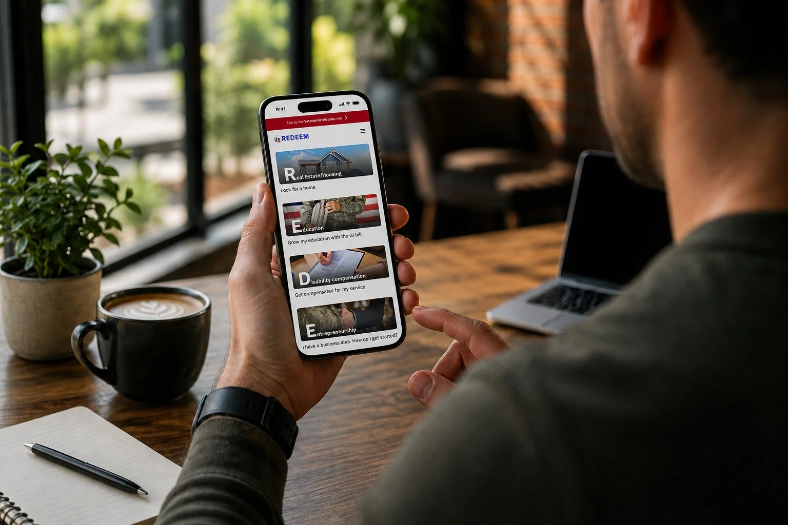 Person holding a smartphone displaying a homepage with categories like Real Estate, Education, Disability compensation, Entrepreneurship, on a wooden table in a cozy café or office setting.