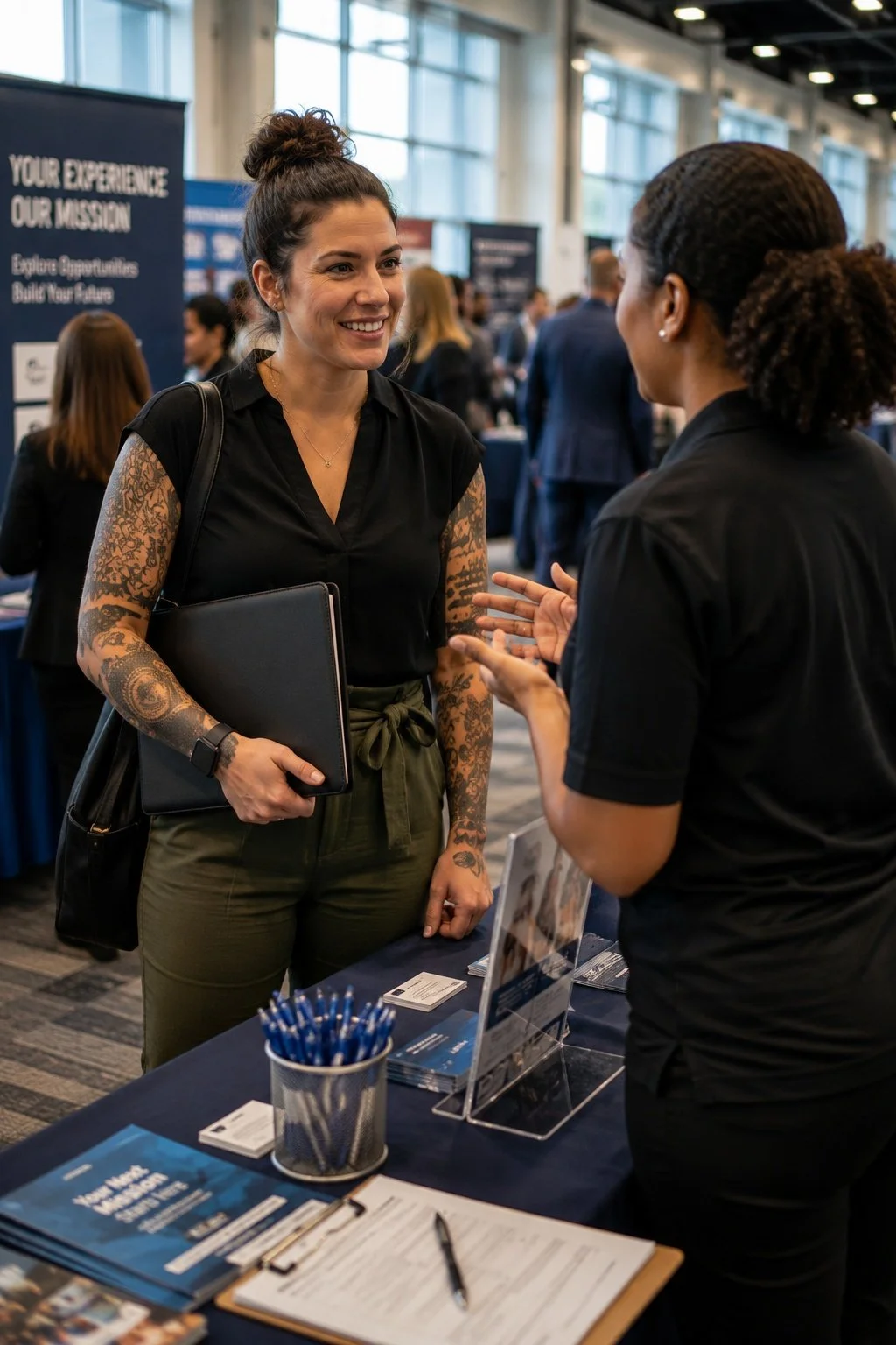 Two women engaged in conversation at a professional event or job fair, with booths and banners in the background.