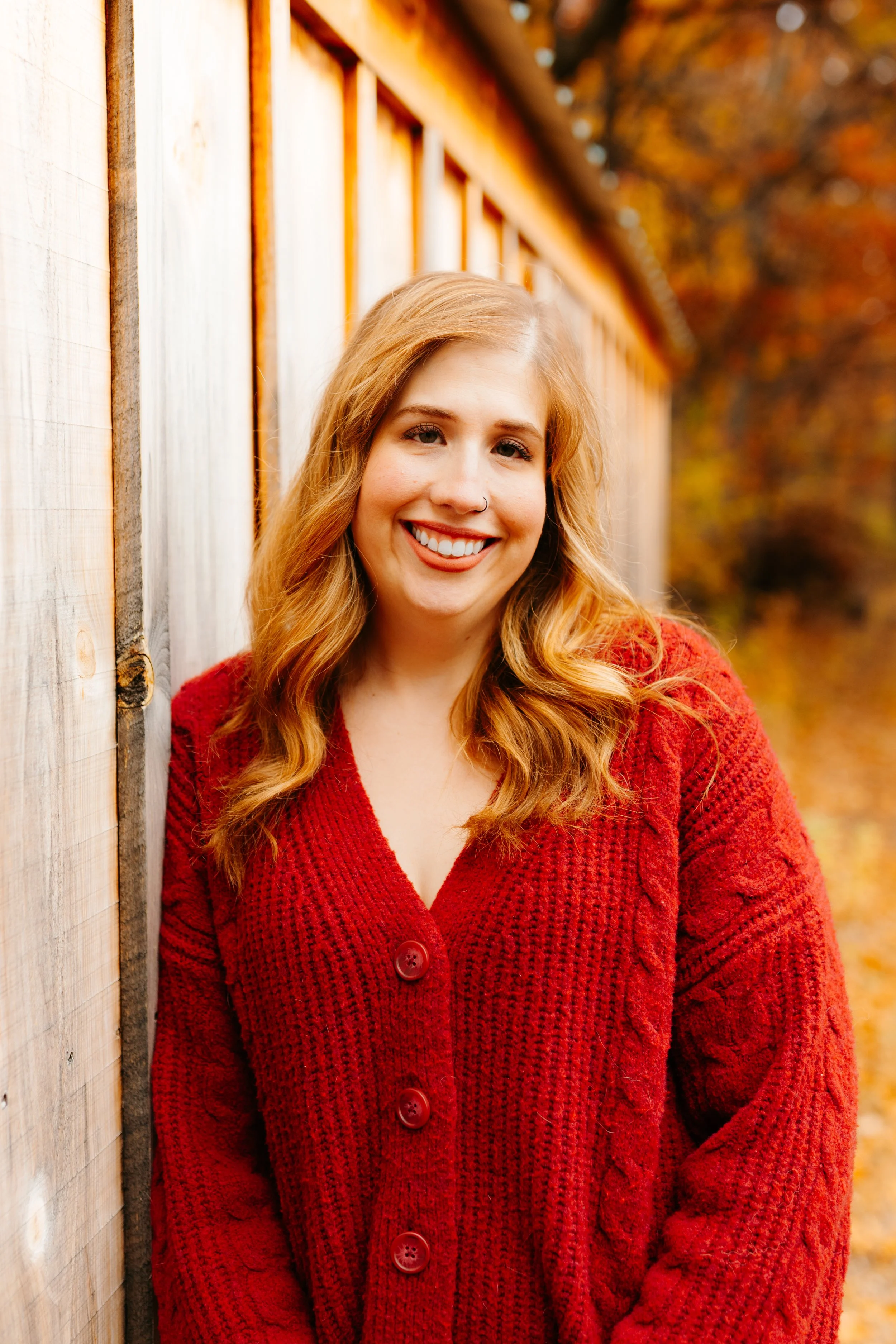 A woman with long wavy red hair, smiling, wearing a red knitted cardigan, standing outdoors near a wooden fence, with autumn-colored trees in the background.